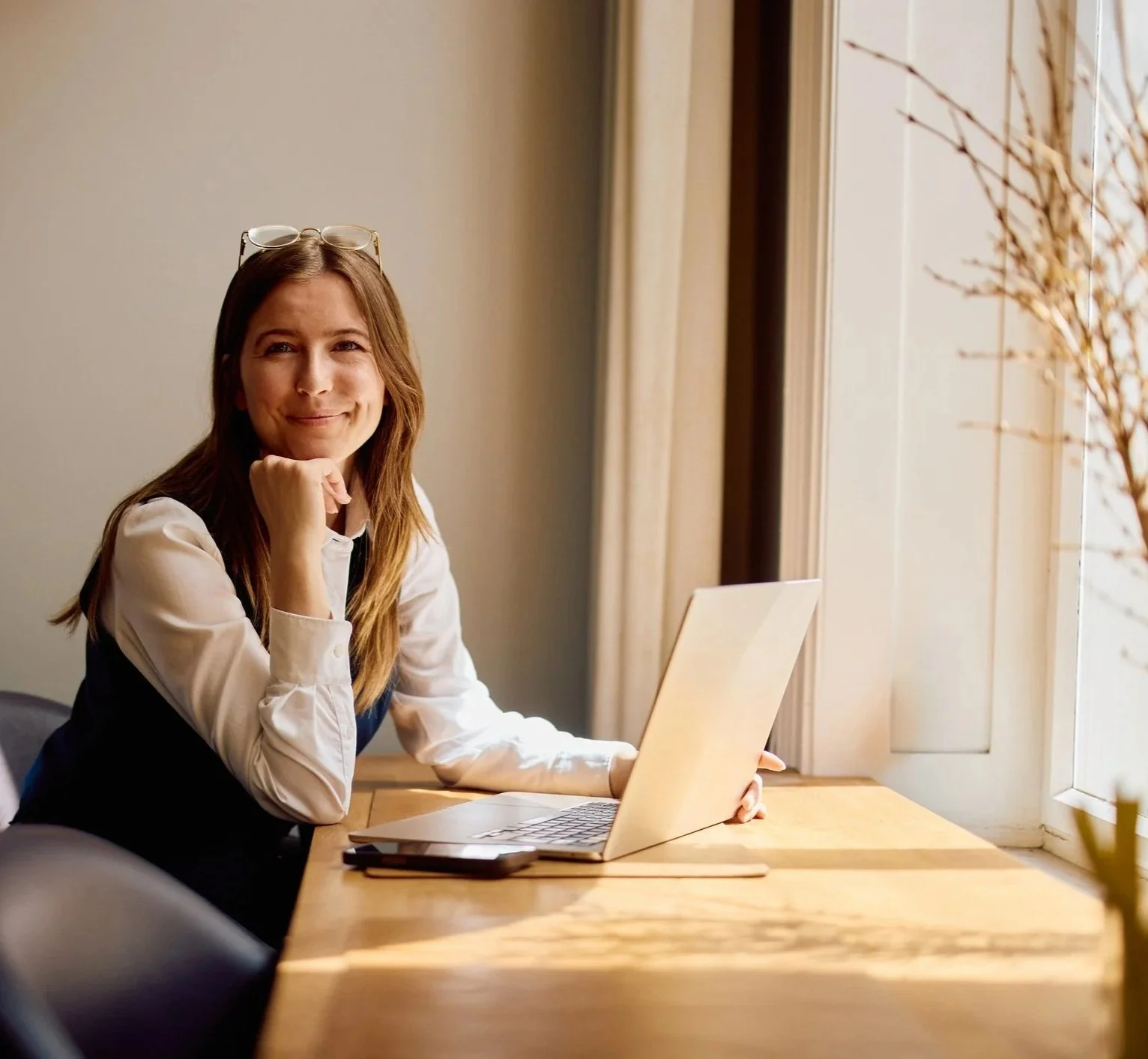 A woman sitting at a wooden table near a window with sunlight, working on a laptop, wearing glasses on her head and smiling.