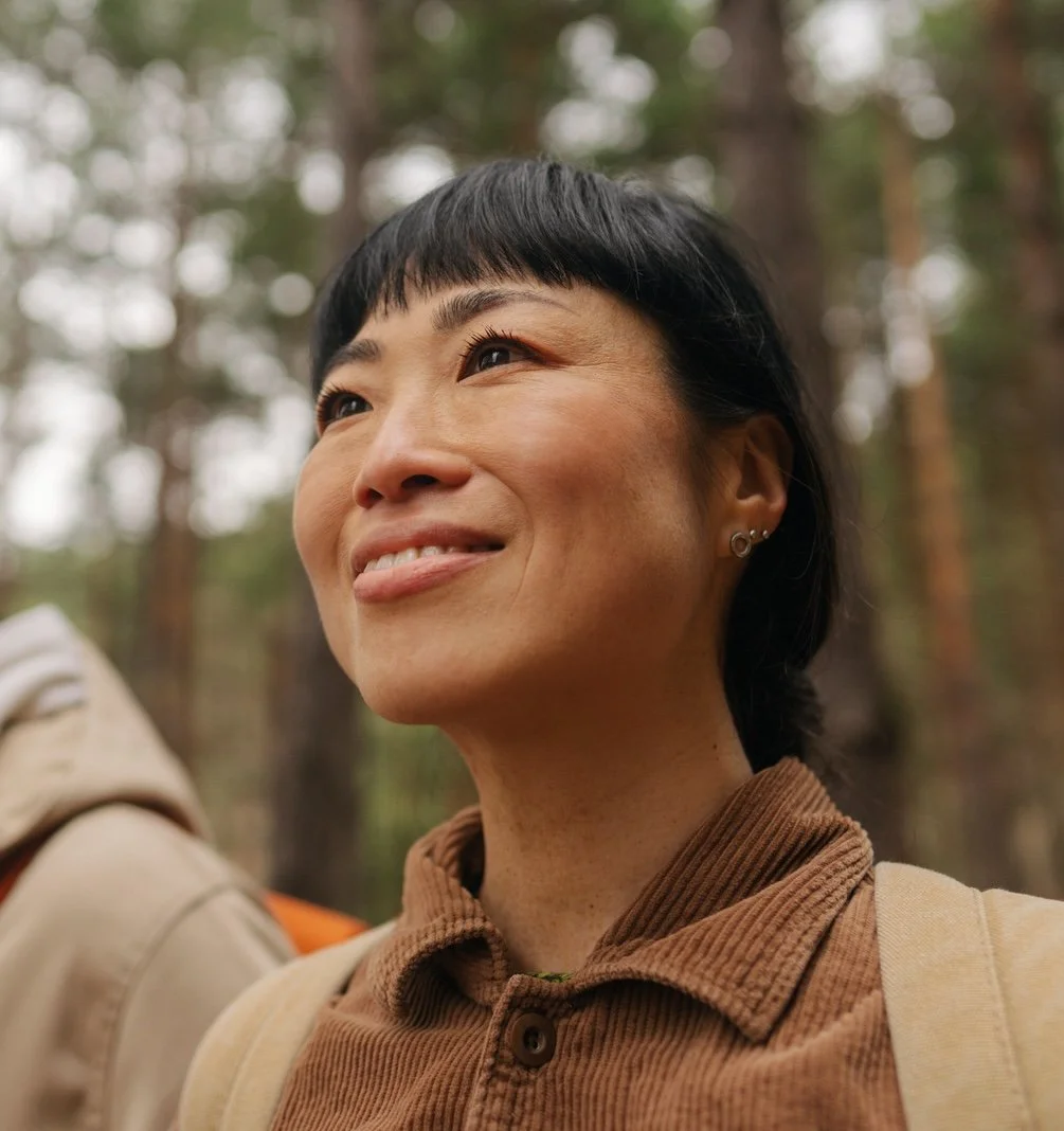 Close-up of a woman with short black hair and earrings smiling outdoors in a wooded area.