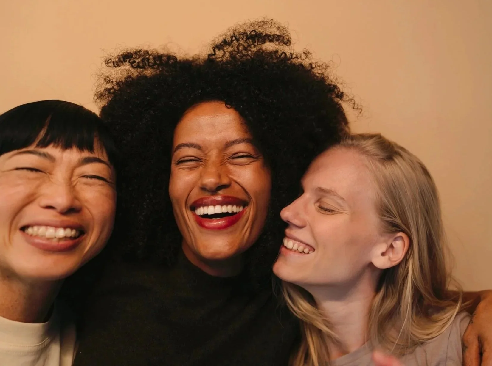 Three women smiling and laughing together in close-up, showing joyful expressions.
