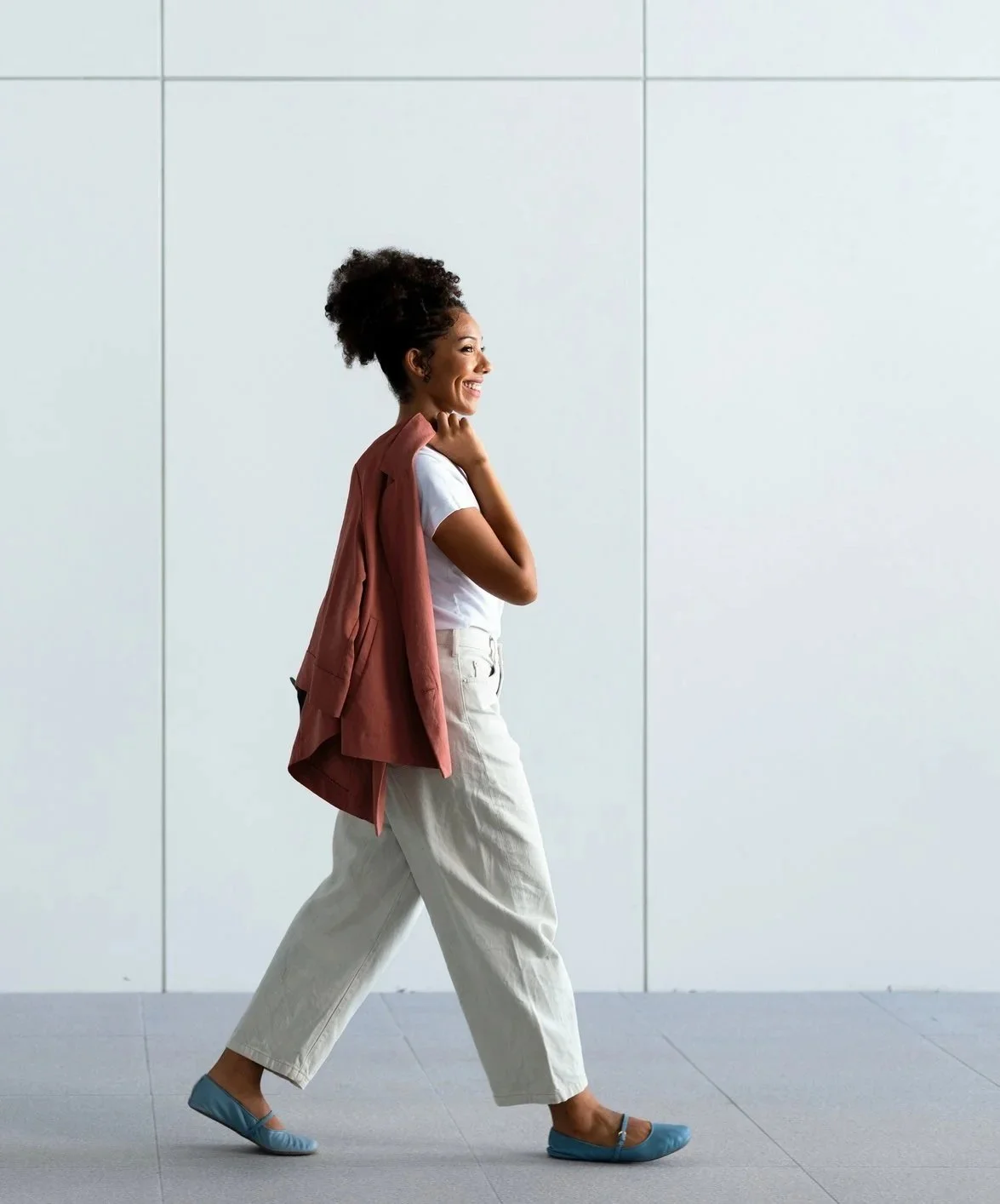 A woman walking in profile with a smile, wearing a white t-shirt, loose white pants, blue flats, and carrying a pink blazer over her shoulder, against a plain white wall.