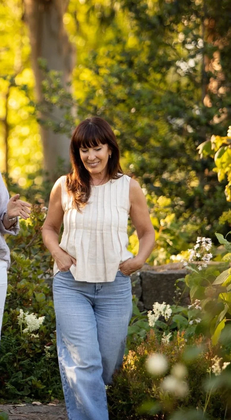 Woman with long dark hair smiling, wearing a sleeveless beige top and light blue jeans, walking through a lush garden with green foliage and white flowers during daylight.