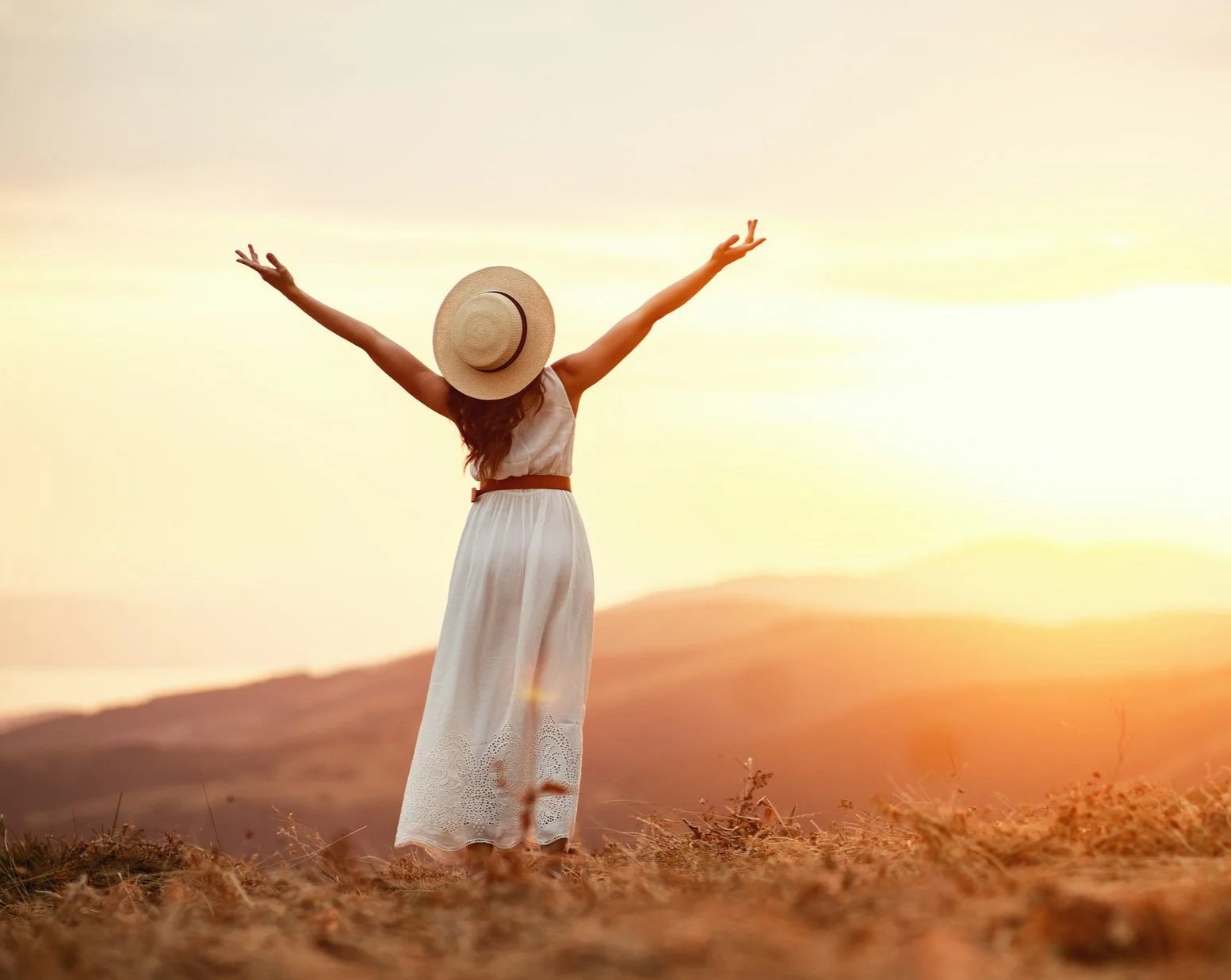 A woman wearing a hat and white dress standing outdoors with arms raised during sunset.
