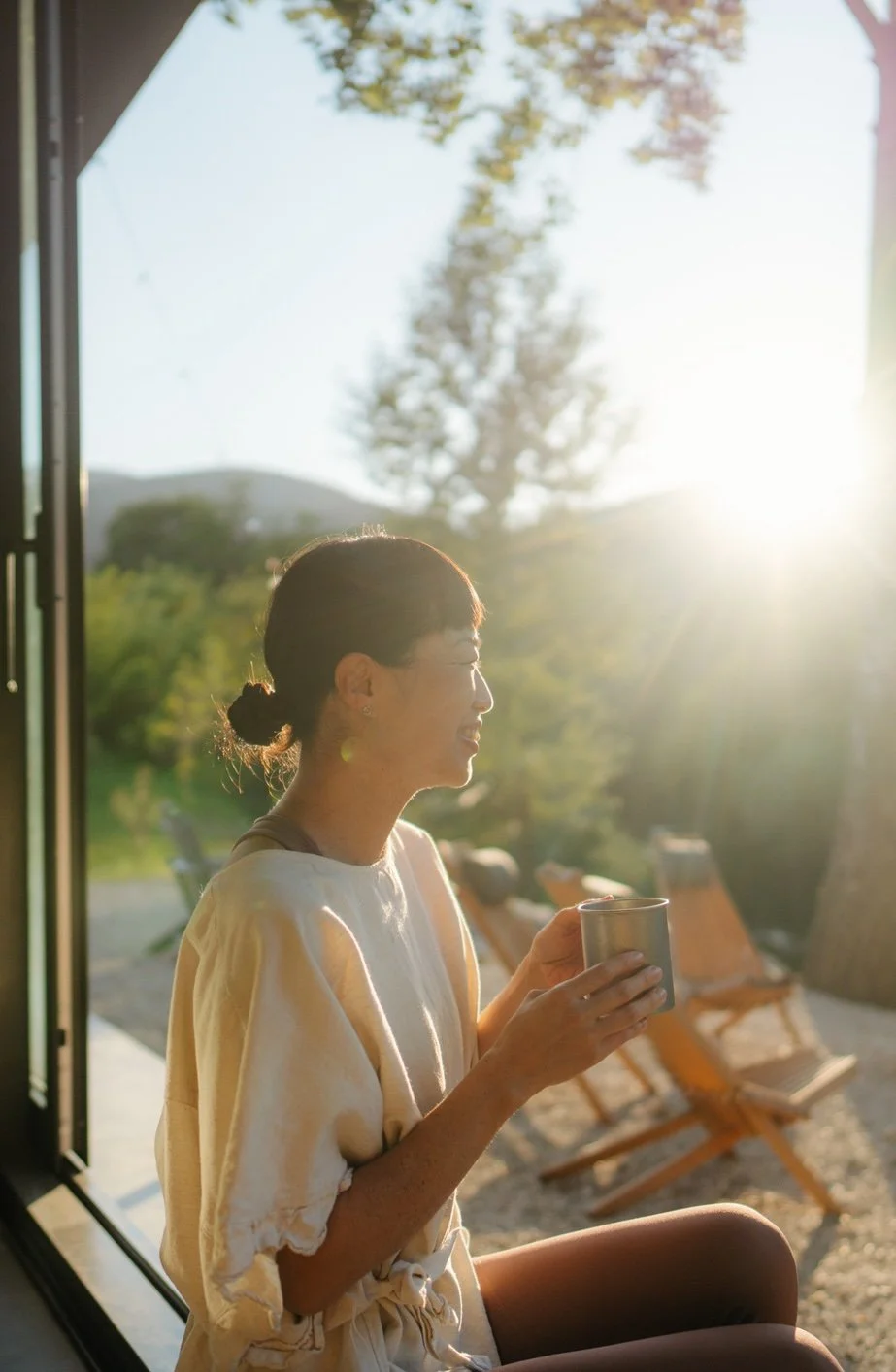 A woman sitting at the threshold of a sliding glass door, holding a mug, enjoying the sunlight during sunset or sunrise on the porch with wooden chairs and a tree in the background.