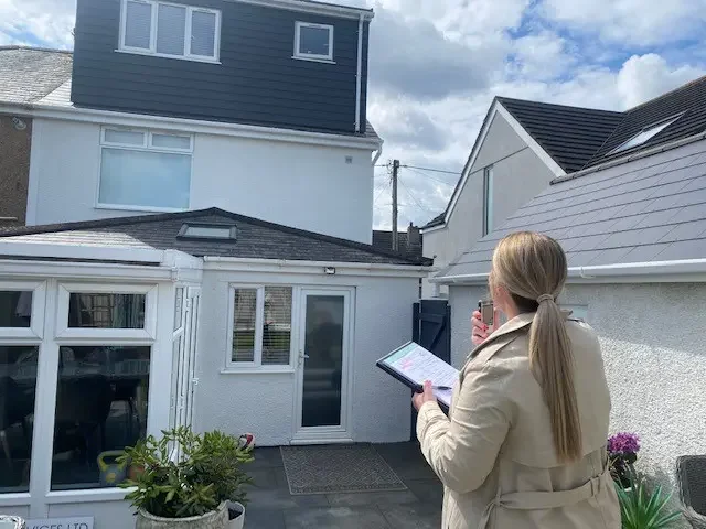 Woman with blonde hair in a ponytail, wearing a beige trench coat, holding a tablet and a pink phone, standing in a backyard with houses and a greenhouse in the background.