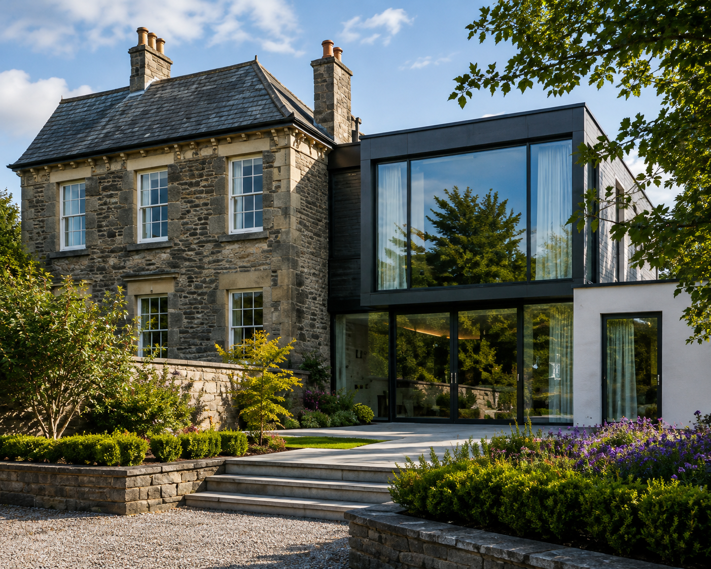 A modern addition to a stone house features large glass windows reflecting greenery, with a landscaped garden and stone steps in the foreground under a blue sky with scattered clouds.