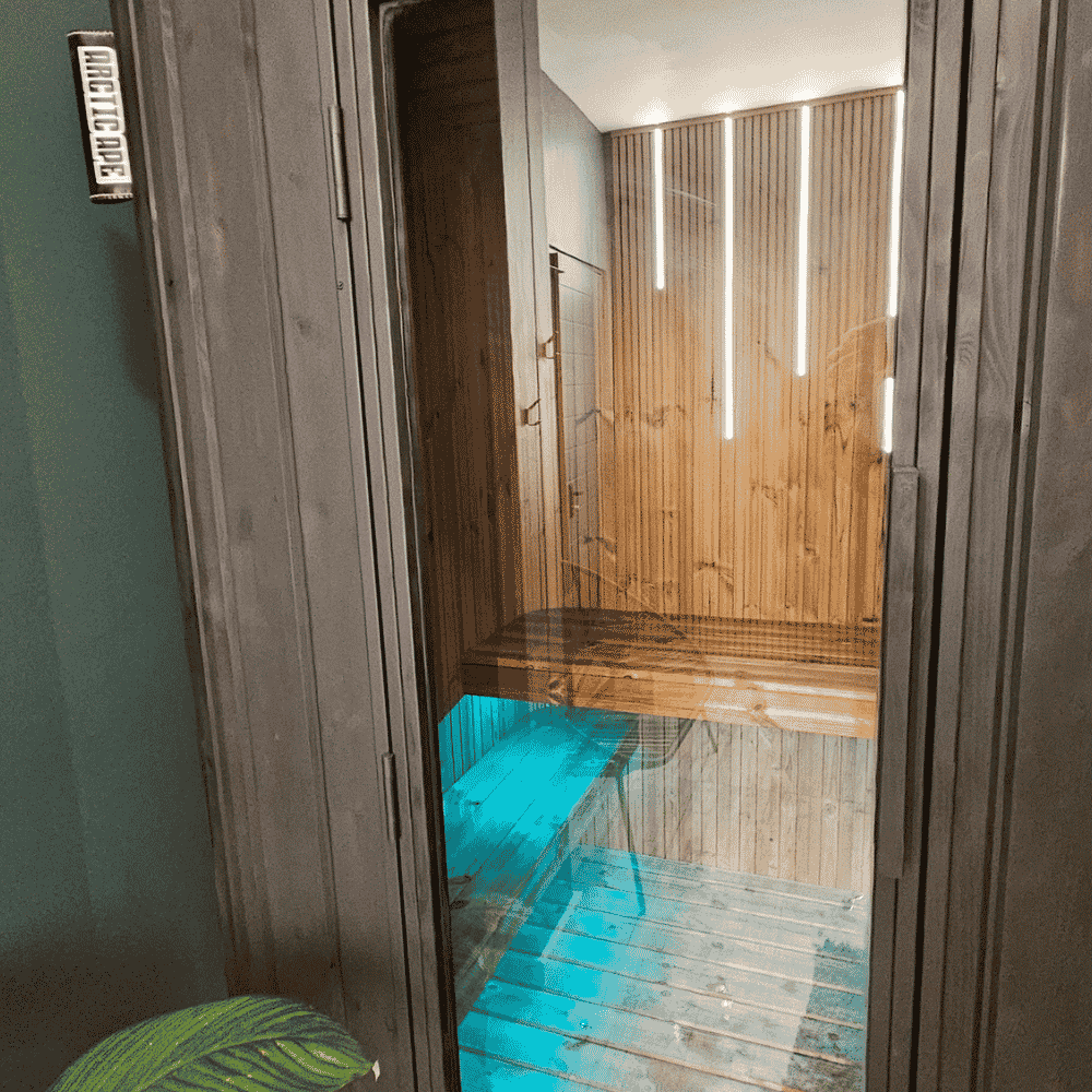 View into a wooden sauna room through a glass door, showing wooden benches and vertical light strips on the wall.