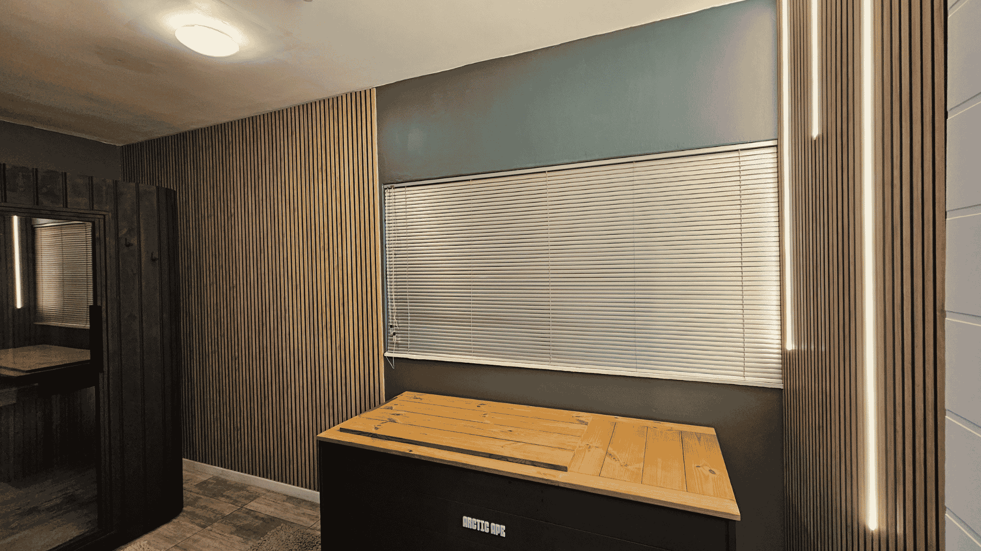 Interior of a room with wood paneling on the walls, closed white horizontal blinds on the window, a beige ceiling with a light fixture, and a wooden table with a black base labeled Arctic Ape.
