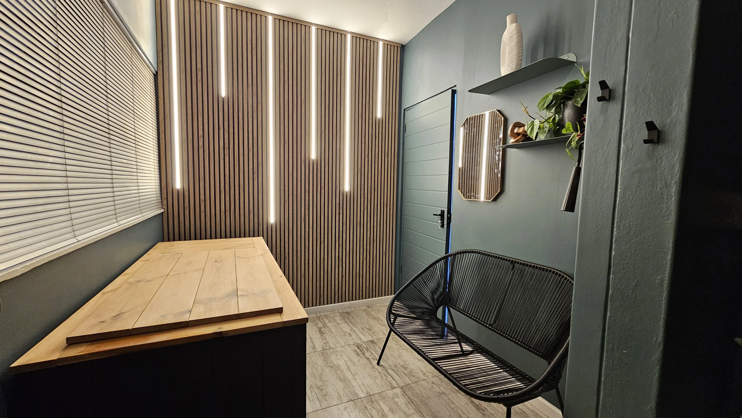 A modern interior room with vertical wood slat wall panels, hanging vertical LED lights, a window with closed blinds, a closed door, a light wooden table, a black wire chair, and a wall-mounted shelf with a decorative item, a potted plant, and a roller shade on an accent wall.