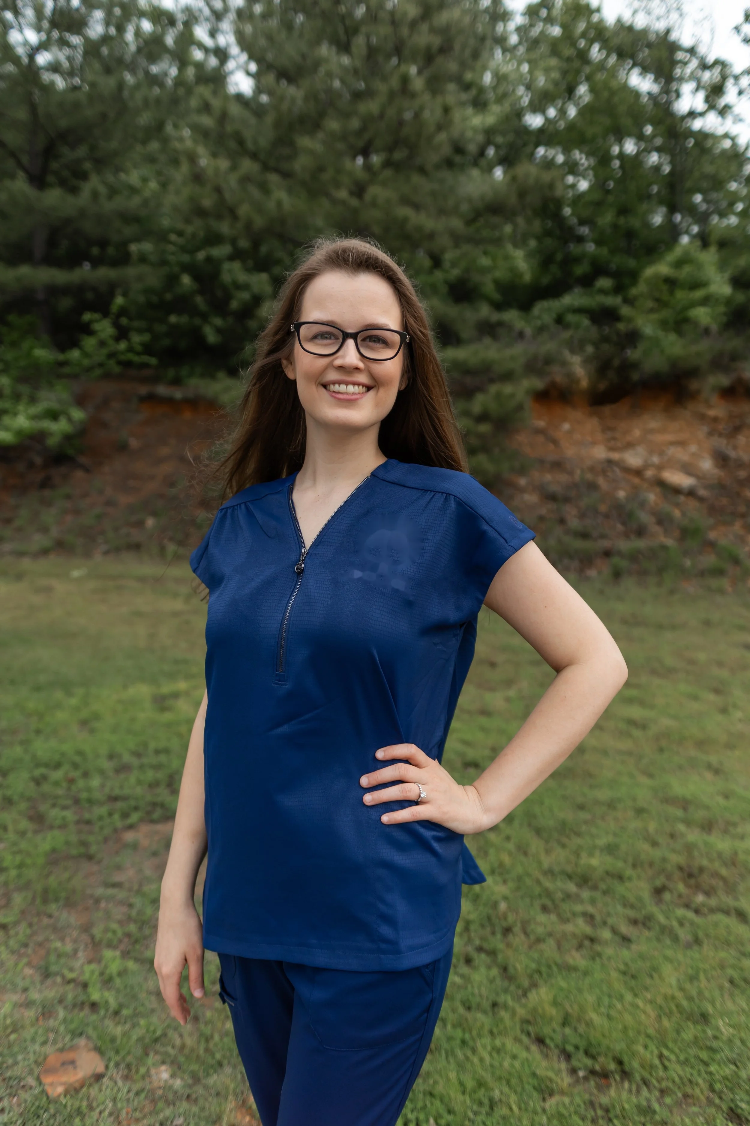 A smiling woman wearing glasses and a blue medical scrub outfit standing outdoors with a hand on her hip, in front of a backdrop of green trees and grass.