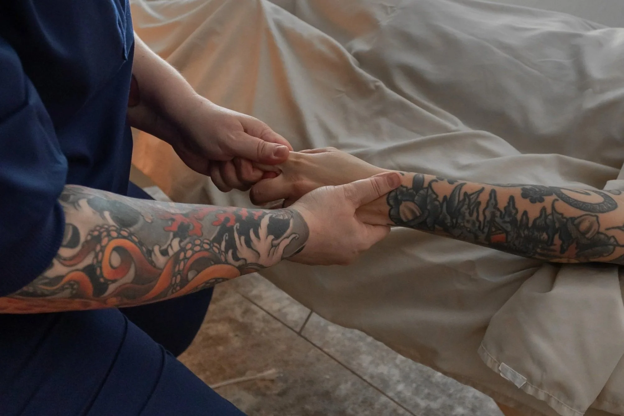 Close-up of tattoos on person's arm, hand grasping another person's wrist, in a setting with a beige tablecloth and tiled floor.