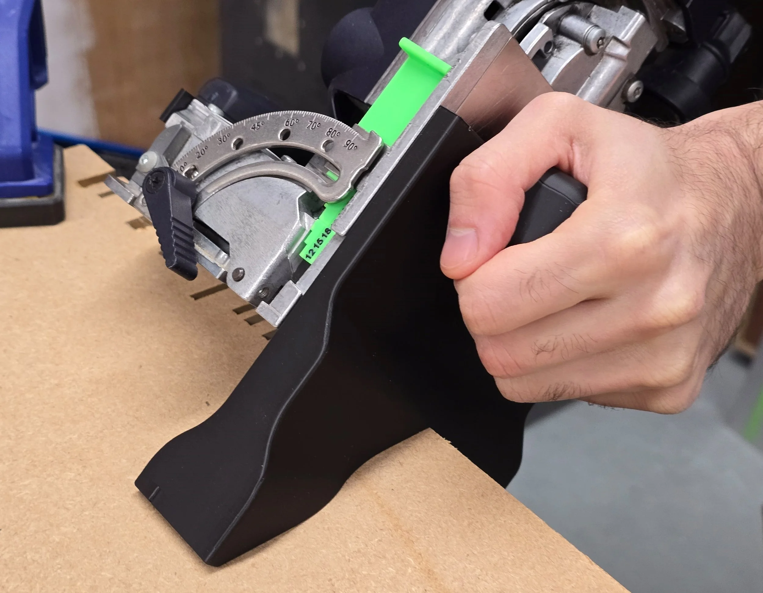 A hand gripping the spline jig's handle, fitted to a Festool Domino DF 500, positioned on the corner edge of an MDF board