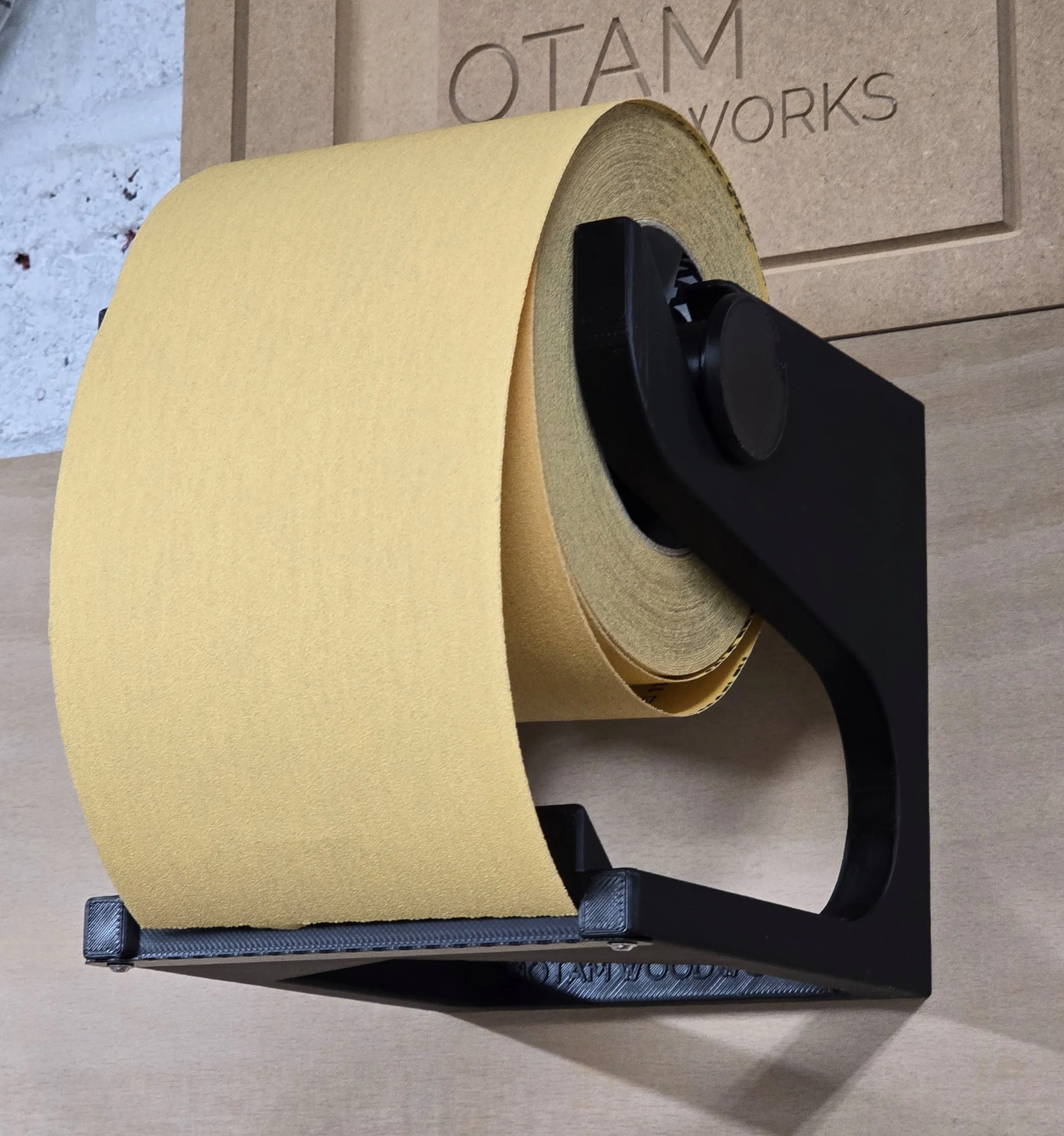 A roll of sandpaper loaded into the Motam Woodworks sandpaper roll storage bracket, mounted against a branded workshop backdrop