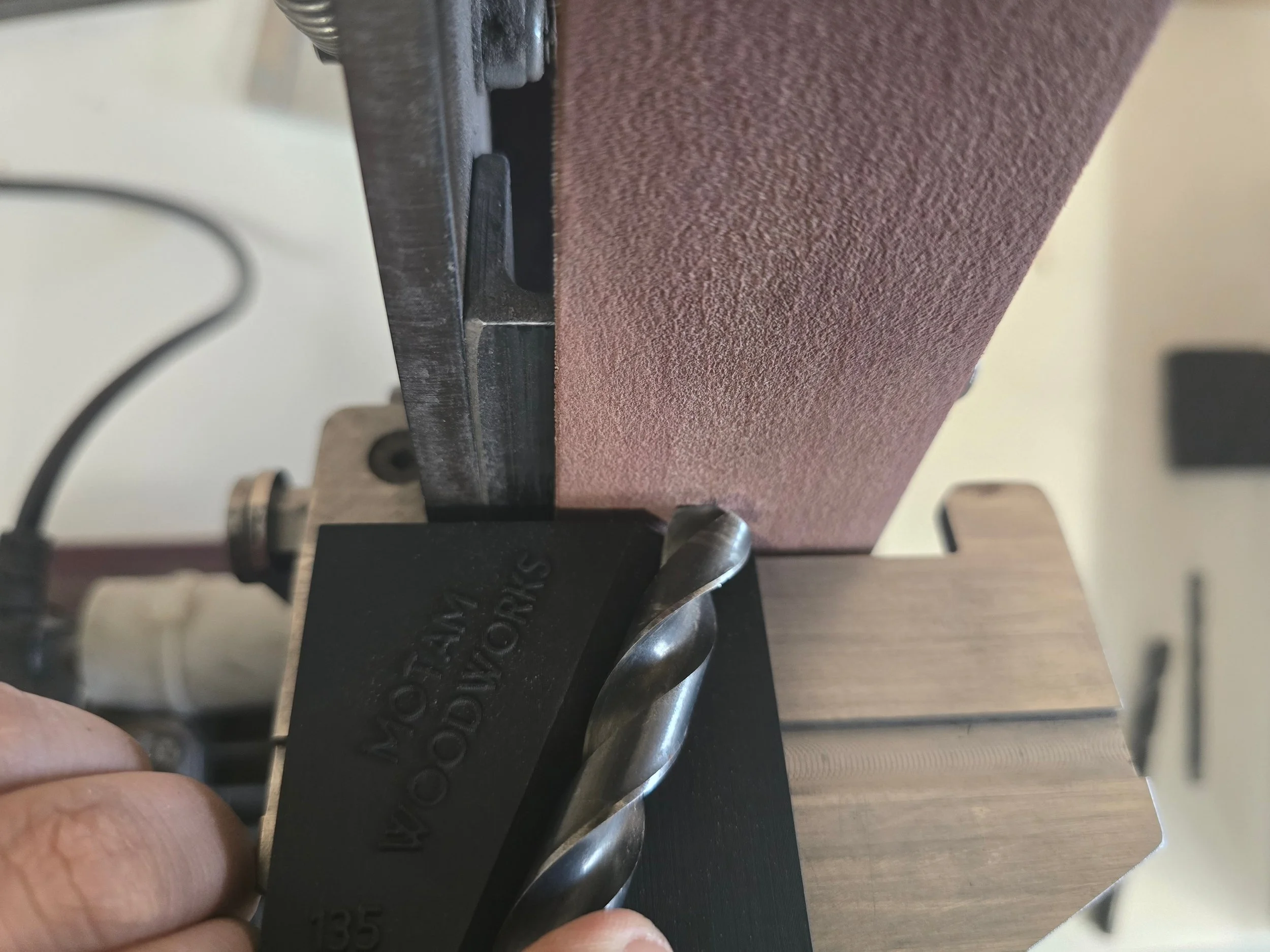 A drill bit held against the sanding belt on the Robert Sorby ProEdge system, guided by the Motam woodworks drill bit jig