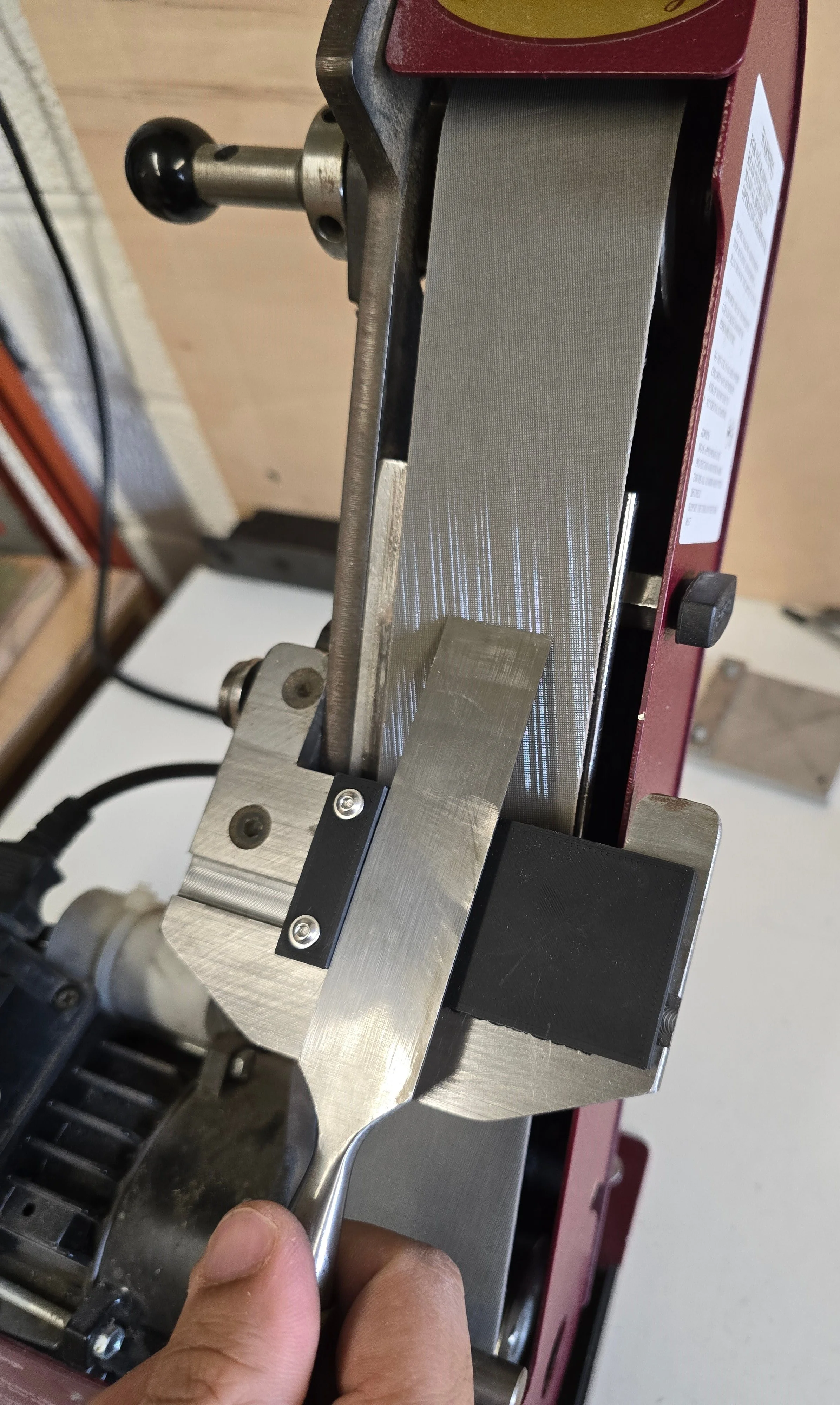 Close-up of the Motam Woodworks square guide jig mounted on the Robert Sorby ProEdge sharpening machine, holding a chisel blade against the sanding belt
