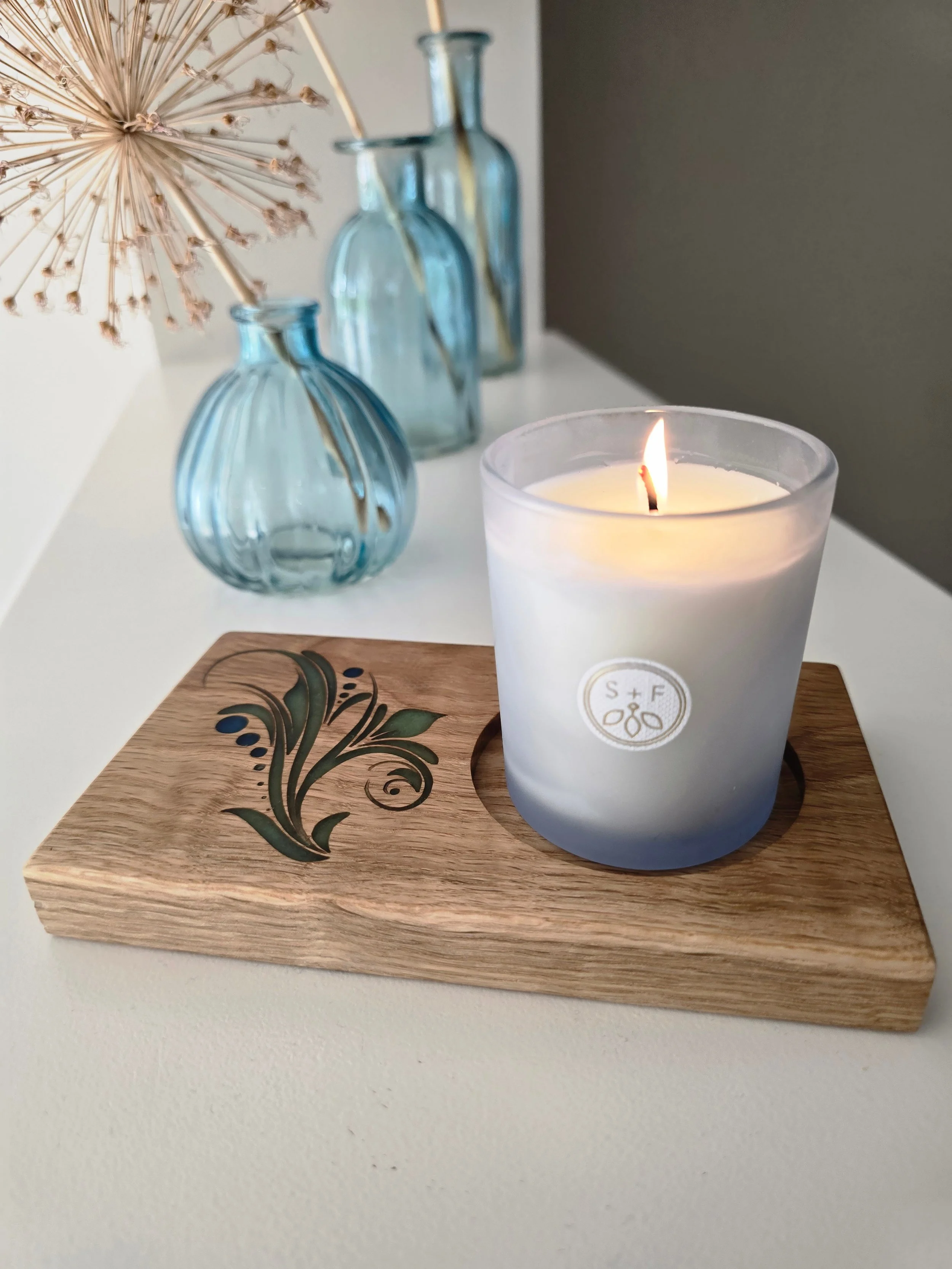 Oak candle holder with green and blue floral resin inlay, styled on a bedside table with dried flowers and glass bud vases, candle lit