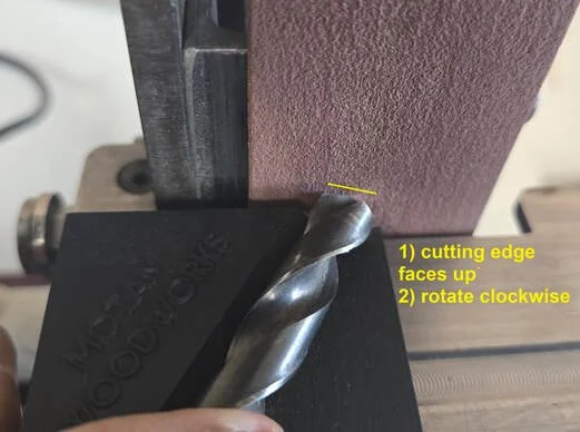 Close-up of a drill bit being used to cut the edge of a piece of wood or material, with instructions to face up and rotate clockwise.