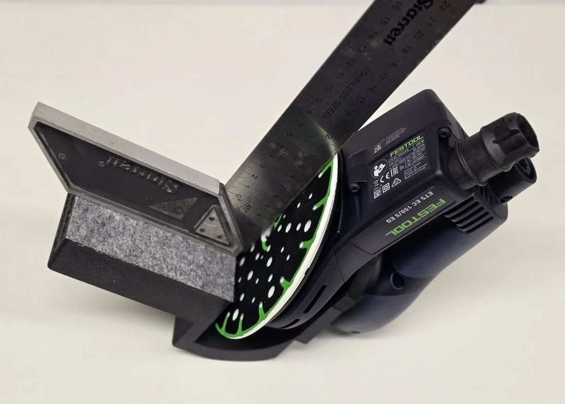 A Festool electric sander with a black handle, a green and black circular sanding pad, a metal ruler, and a small rectangular sanding block on a white surface.