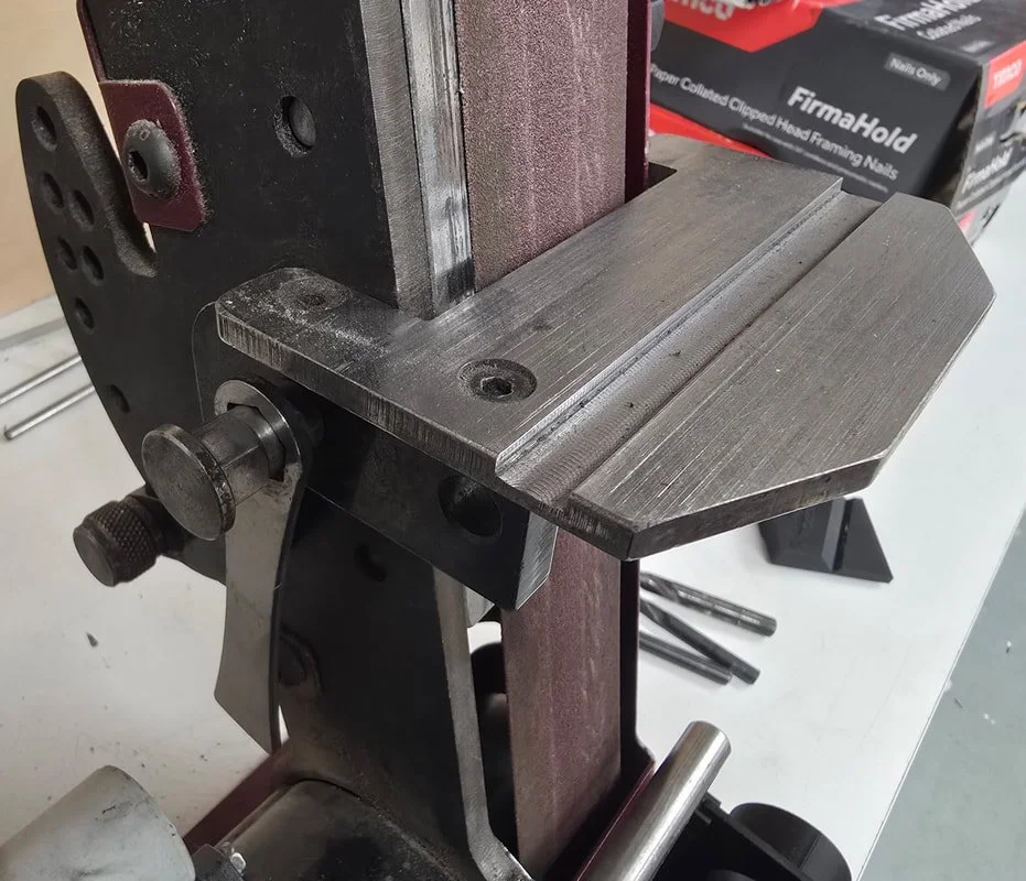 Close-up of a metal woodworking or machining tool with adjustable parts, set on a white work surface, with tool bits and boxes in the background.