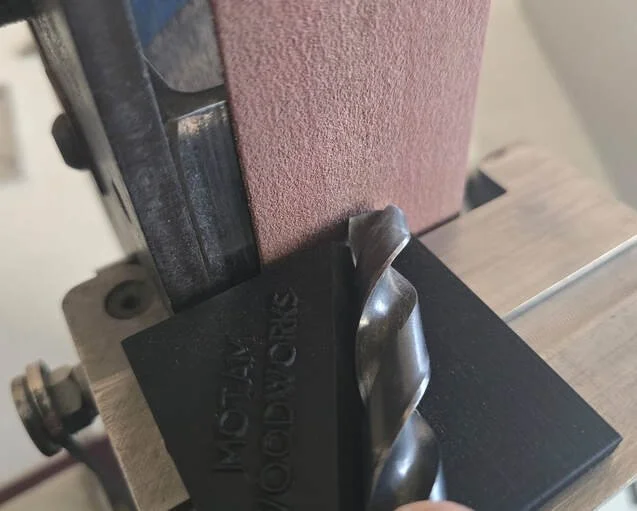 Close-up of a metal drill bit mounted on a drill press with a pink abrasive wheel in the background.