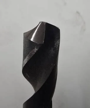 Close-up of a black drill bit with a pointed tip against a plain, light gray background.