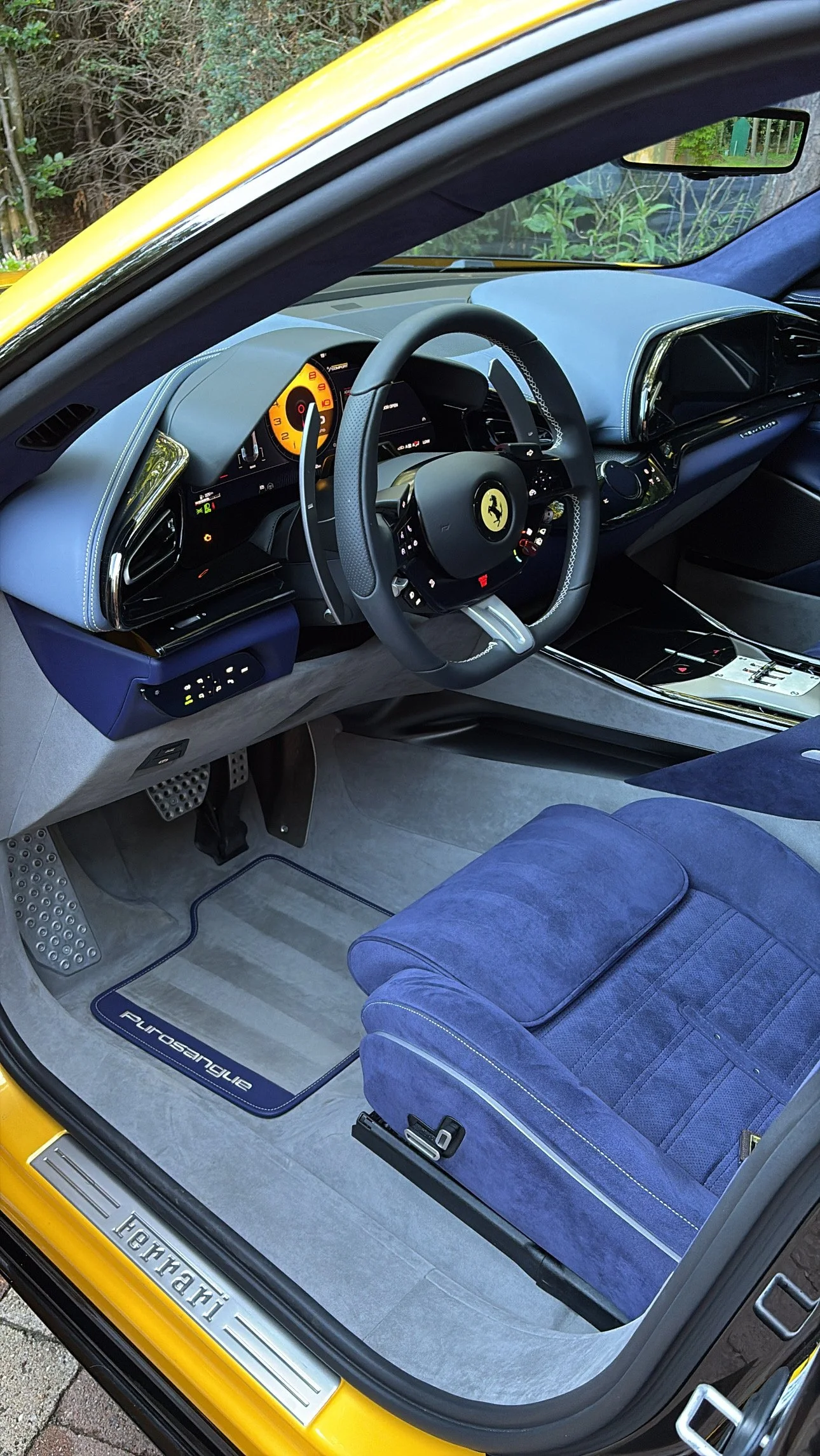 Interieur van een gele Ferrari Purosangue met een zwart stuurwiel, blauw beklede stoelen en een vloer mat met de naam 'Purosangue'.