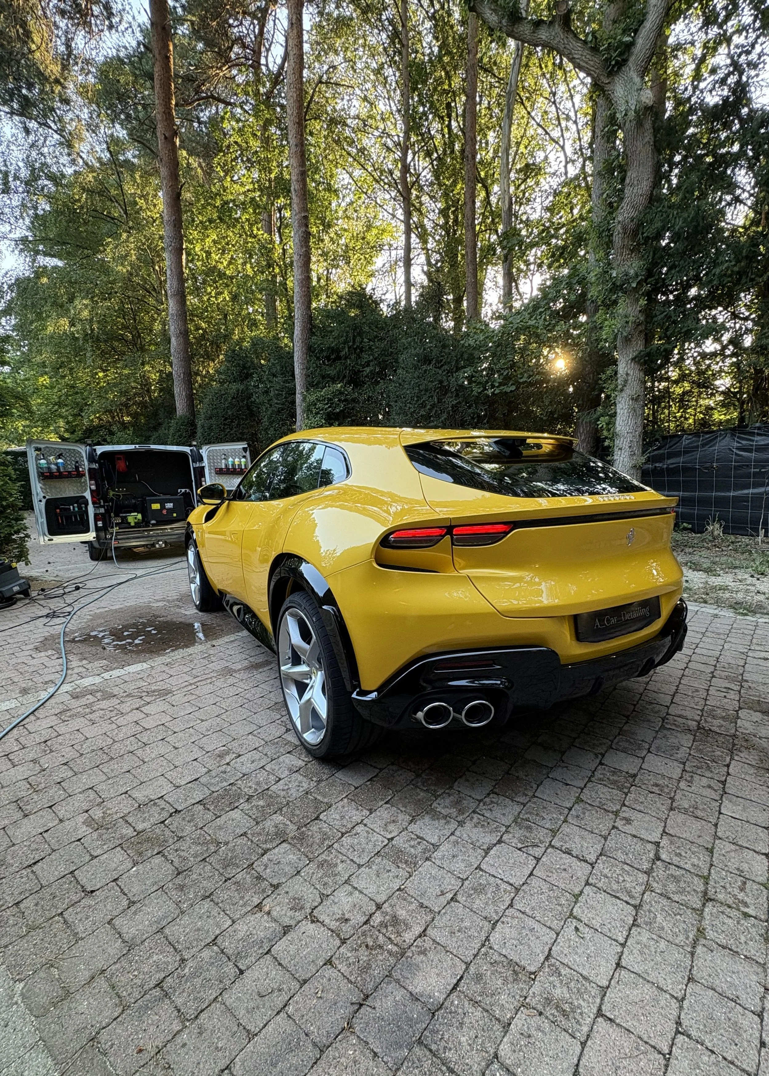 Een felgele sportauto wordt gewassen in een tuin met stenen bestrating, terwijl een mobiele was- of reinigingsstation naast de auto staat.