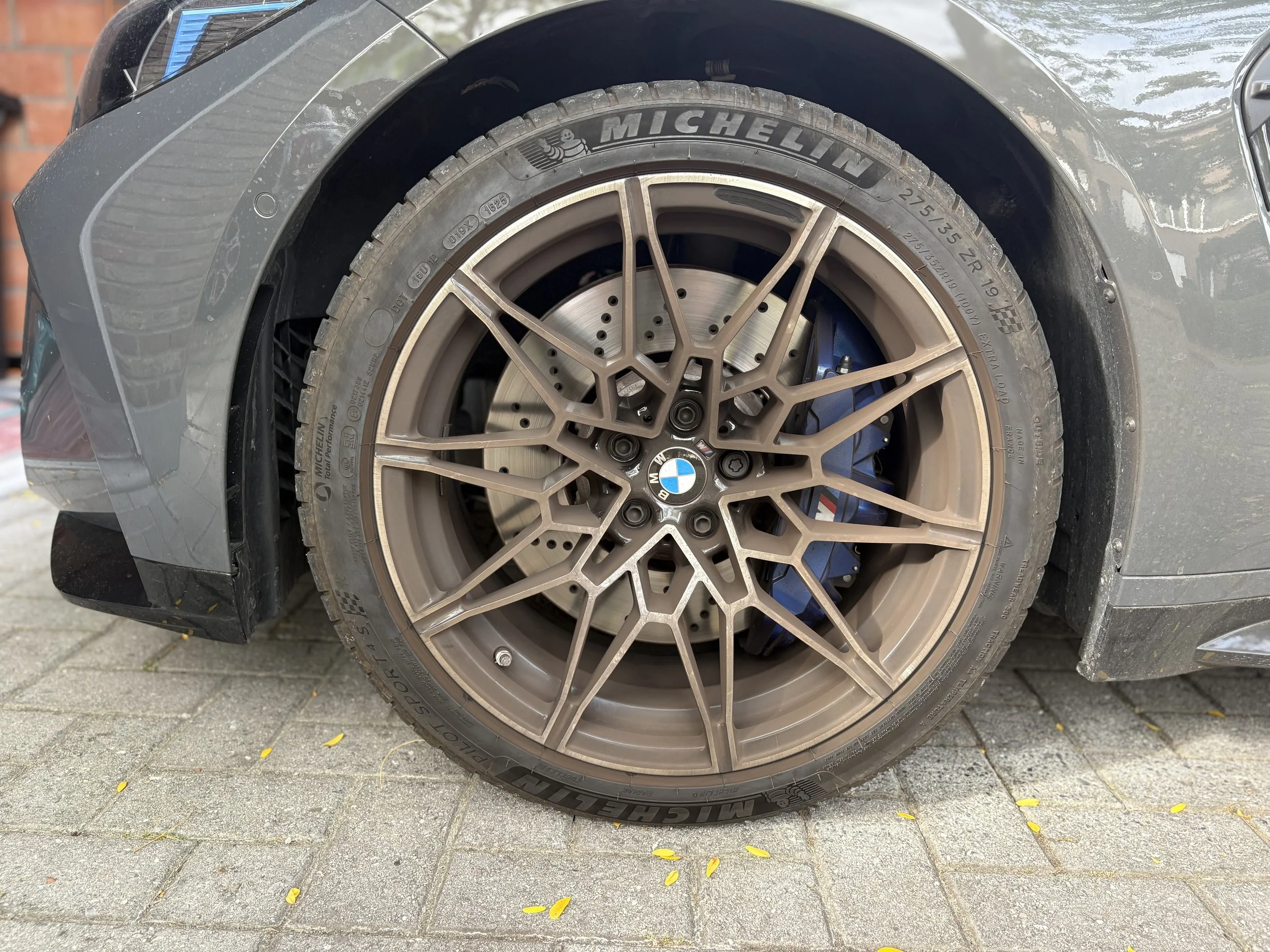 Close-up van een sportwagenwiel met een Michelin-reifen en een bronzen BMW-velg, geparkeerd op een tegelpad met kleine gele blaadjes. De auto is mogelijk een BMW M-model, zichtbaar aan het blauwe remklauw.