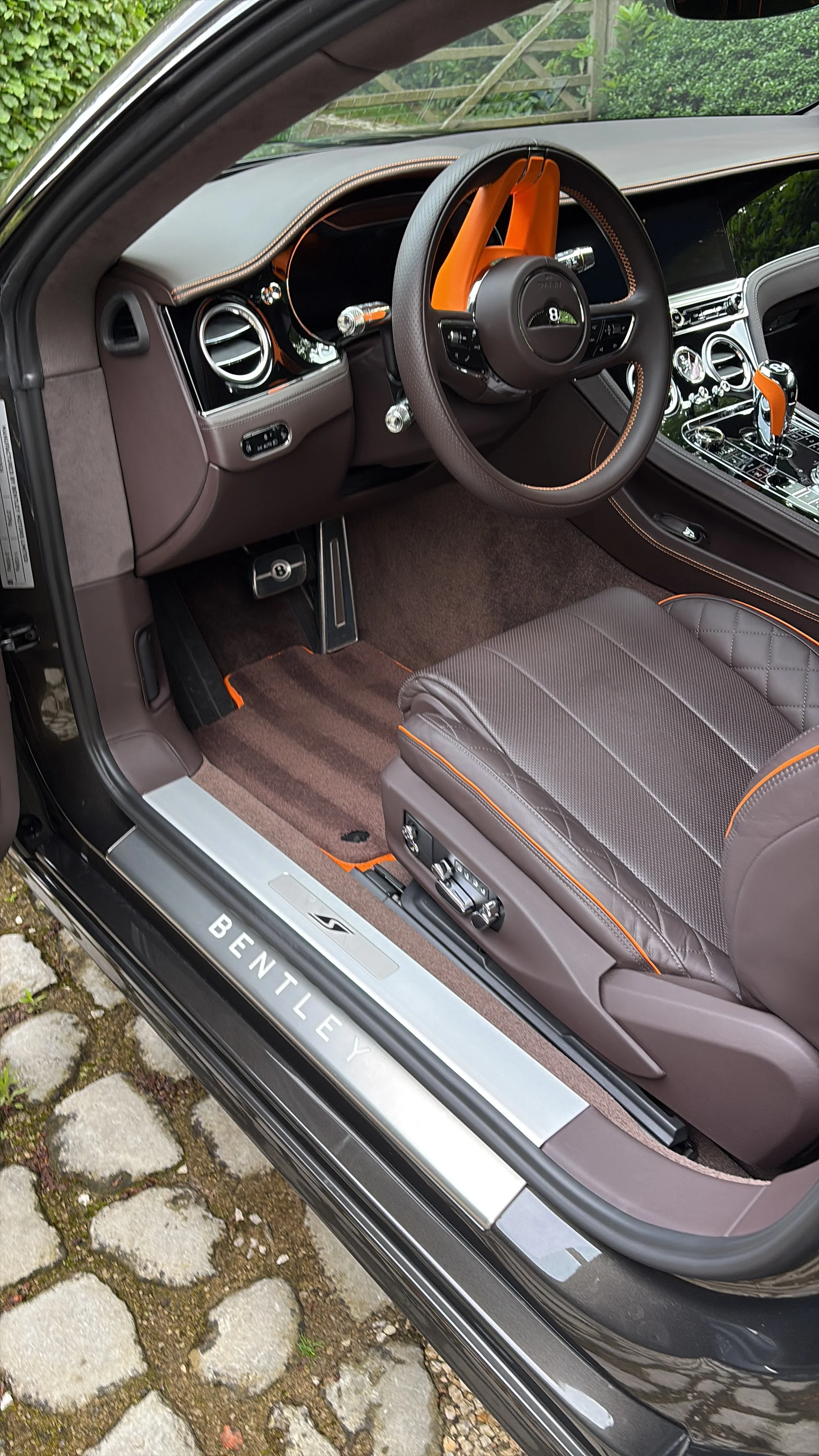 Interieur van een luxe auto, met lederen stoelen, een lederen stuurwiel met oranje accent, en een digitaal dashboard. De deur heeft een metalen rand met 'BENTLEY' erop.