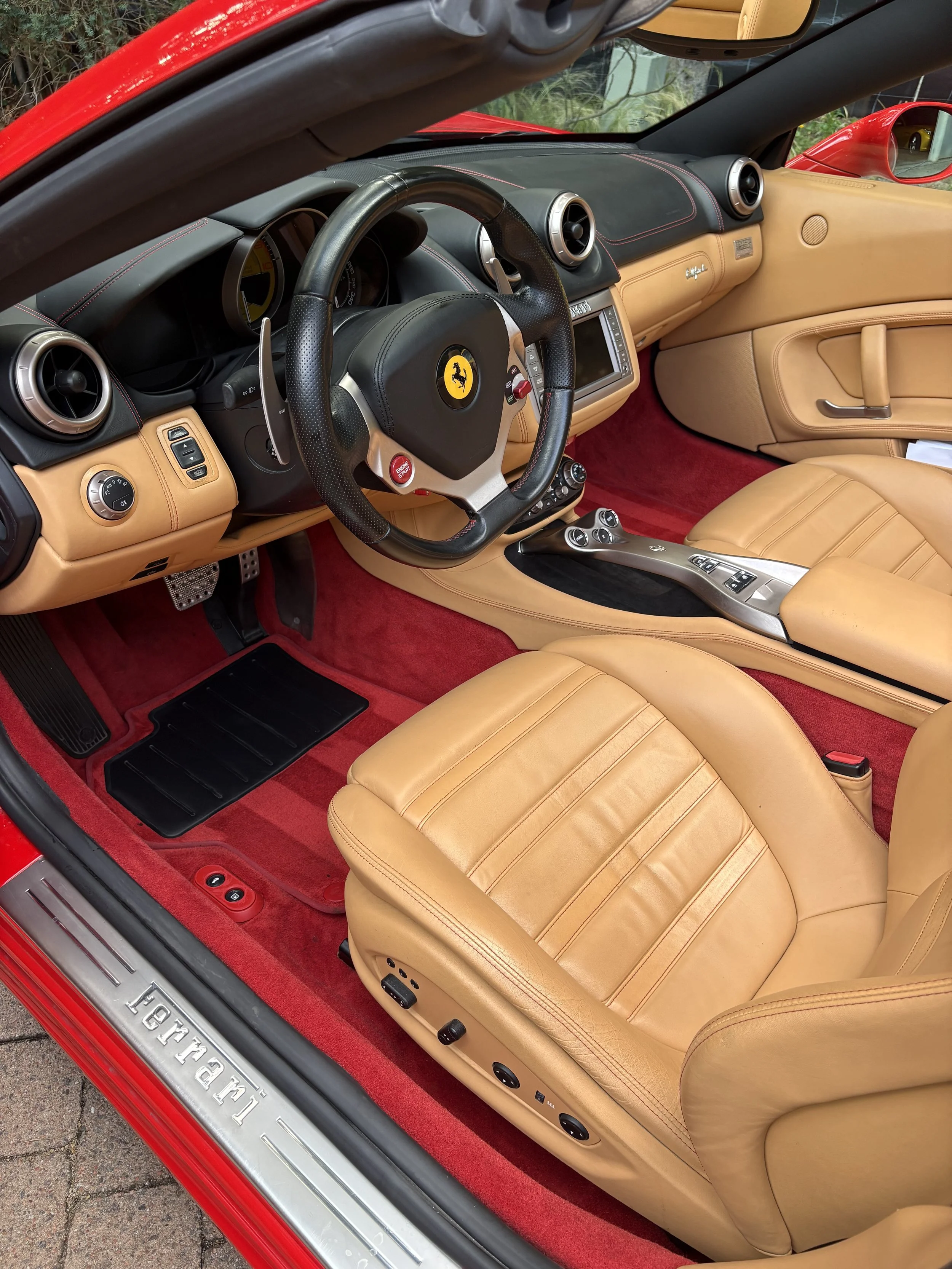 Interieur van een rode Ferrari sportwagen met beige leren stoelen, zwart stuurwiel met Ferrari-embleem, en rode vloerbedekking.