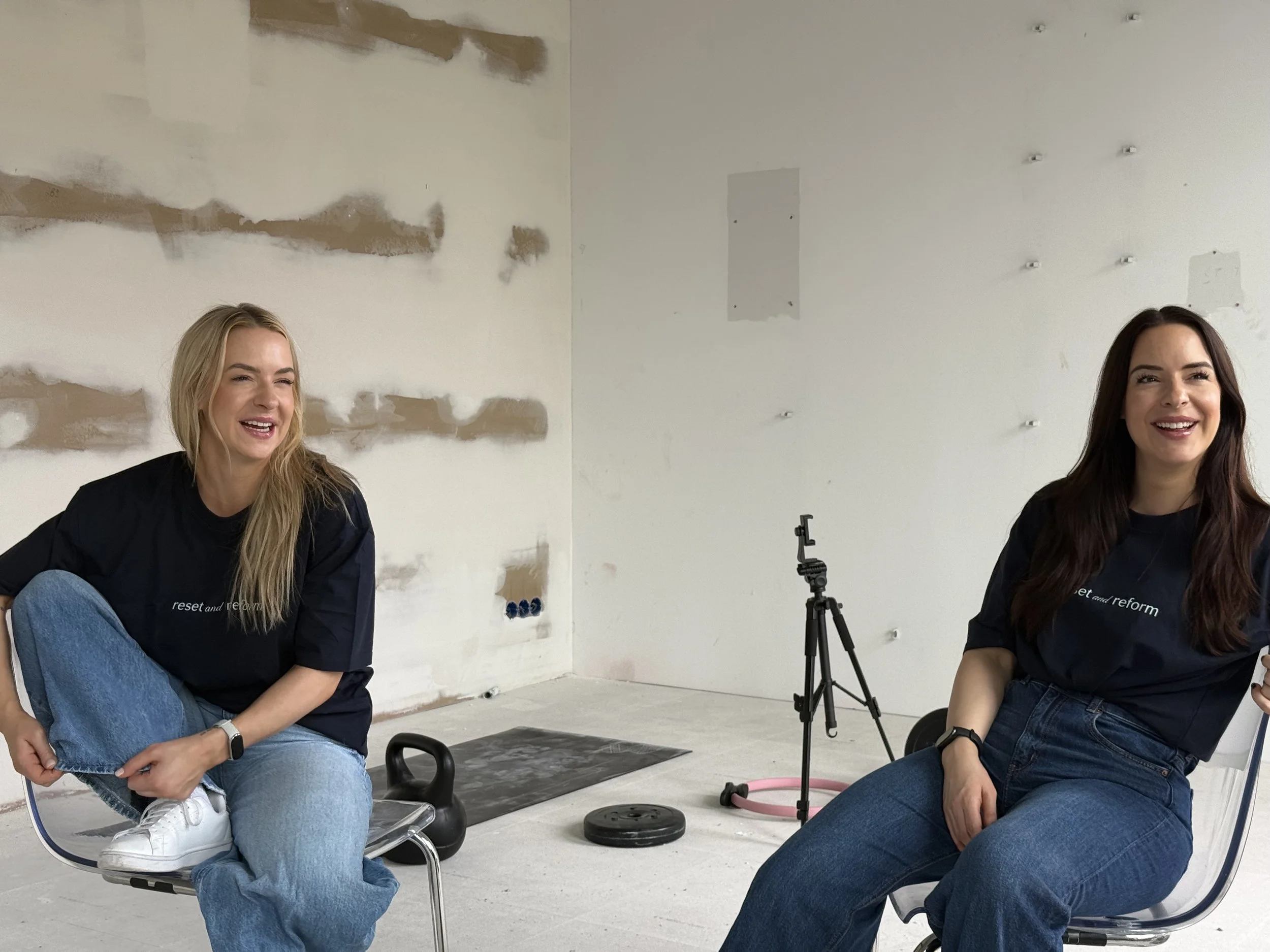 Zwei lachende Frauen sitzen in einem Raum mit unvollständiger Wandverkleidung. Eine trägt ein schwarzes T-Shirt, die andere ein dunkles Oberteil. Es gibt einen schwarzen Gymnastikball, ein Stativ und ein Rollhockey. Beide Frauen wirken entspannt.