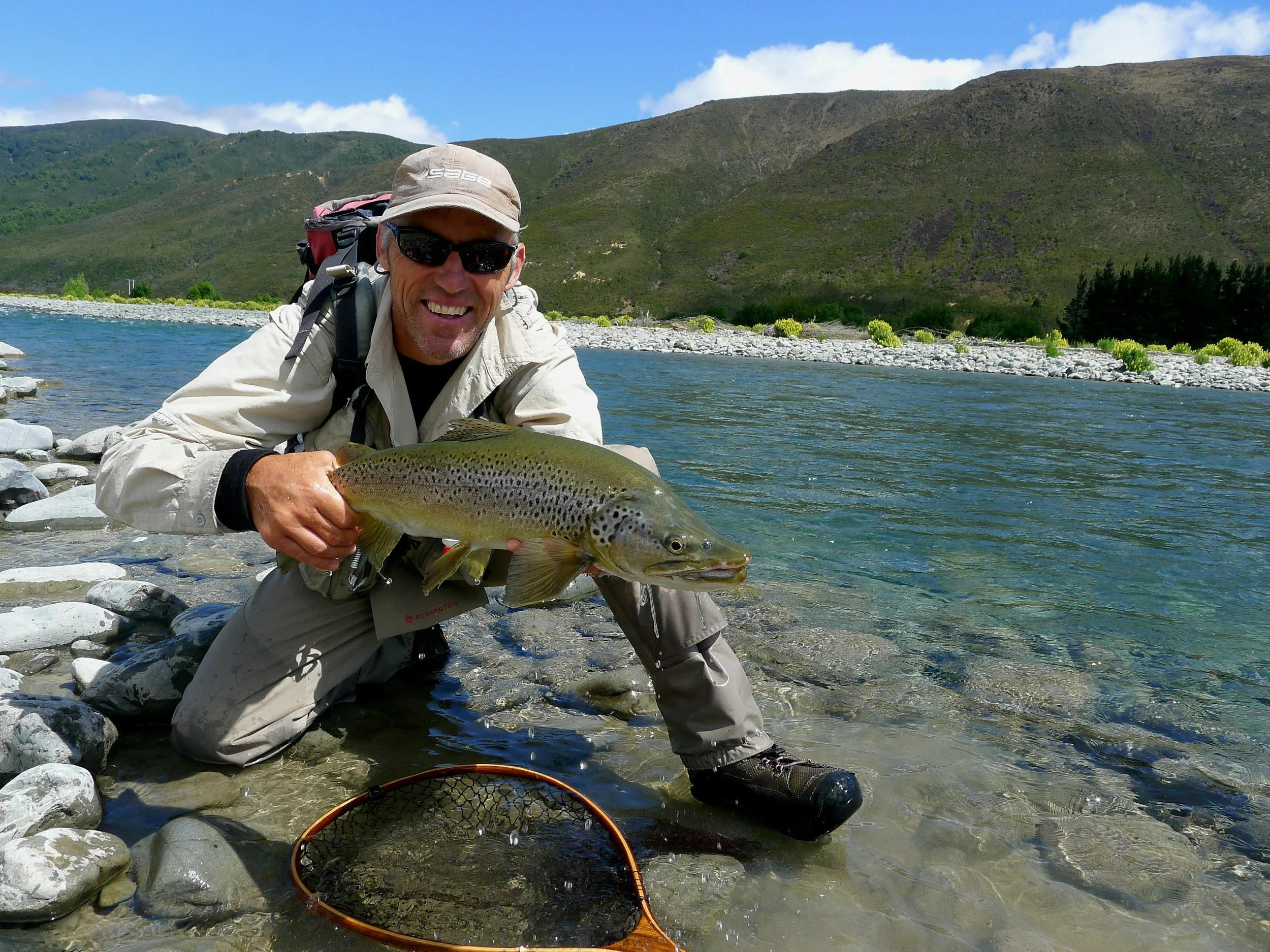 A man with a hat and sunglasses kneeling in a river, holding a large fish he caught, with mountains and a blue sky in the background.