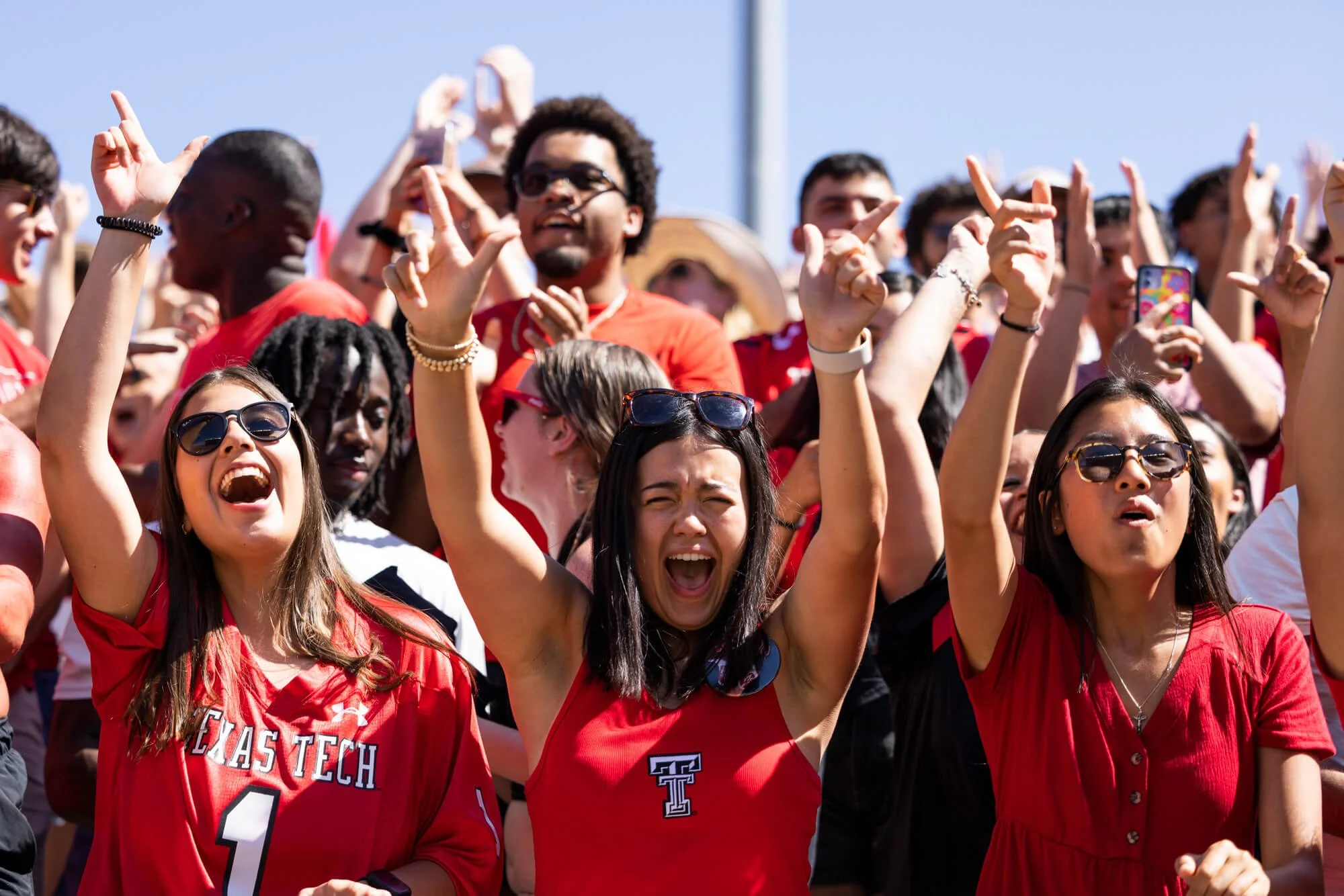 A large group of diverse people wearing red Texas Tech shirts at an outdoor event, some raising their hands and taking photos, with clear blue sky in the background.