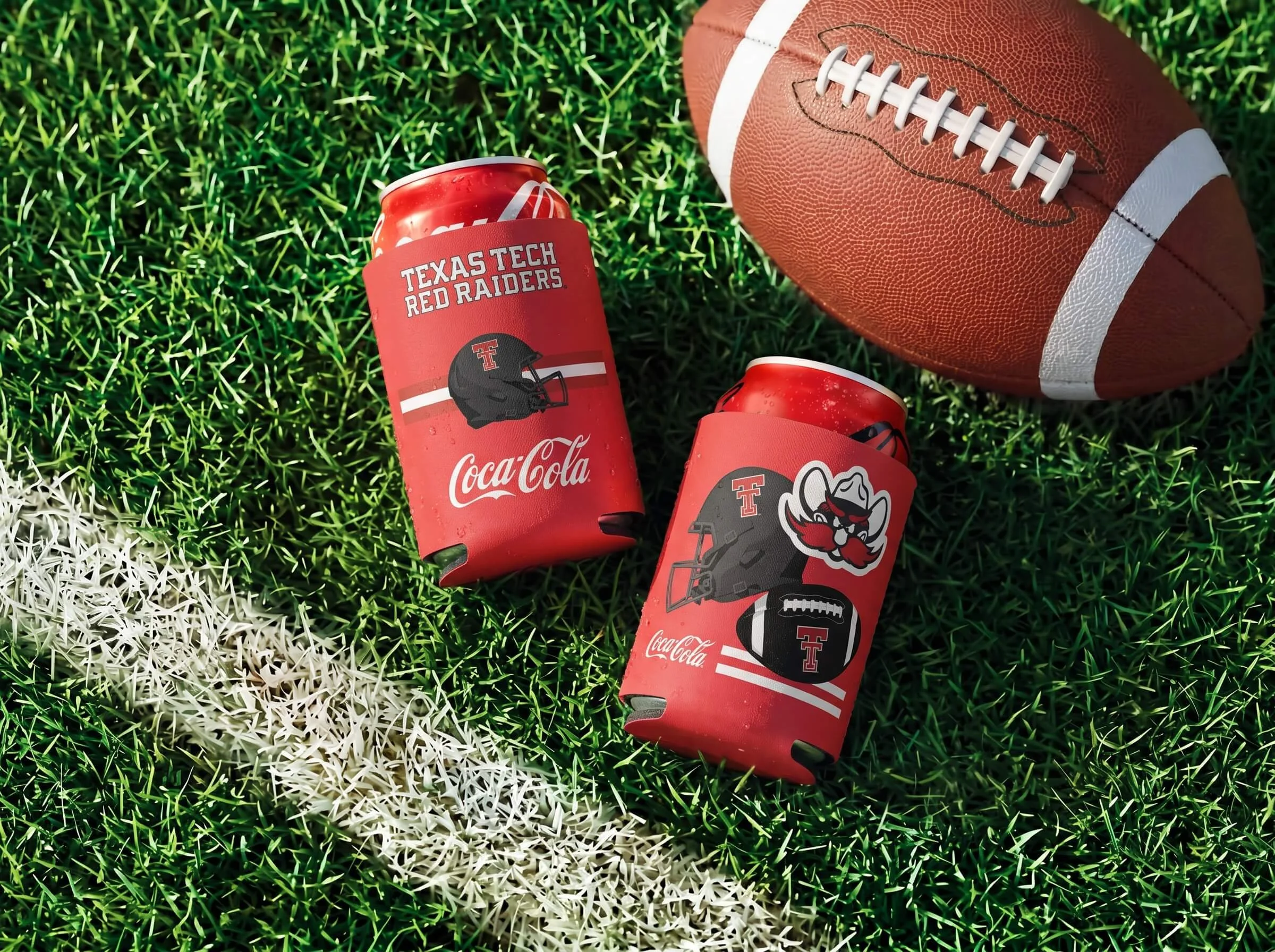 A football on green turf with white and green lines, a red Coca-Cola can with Texas Tech branding, and a red can with a logo of a red Raider mascot.