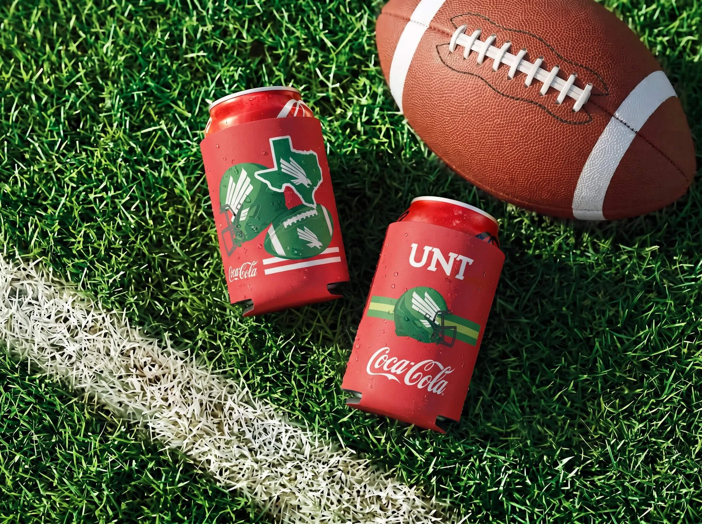 A football lying on a green field next to two cans of Coca-Cola, one with a South Texas themed label and the other with the University of North Texas logo.