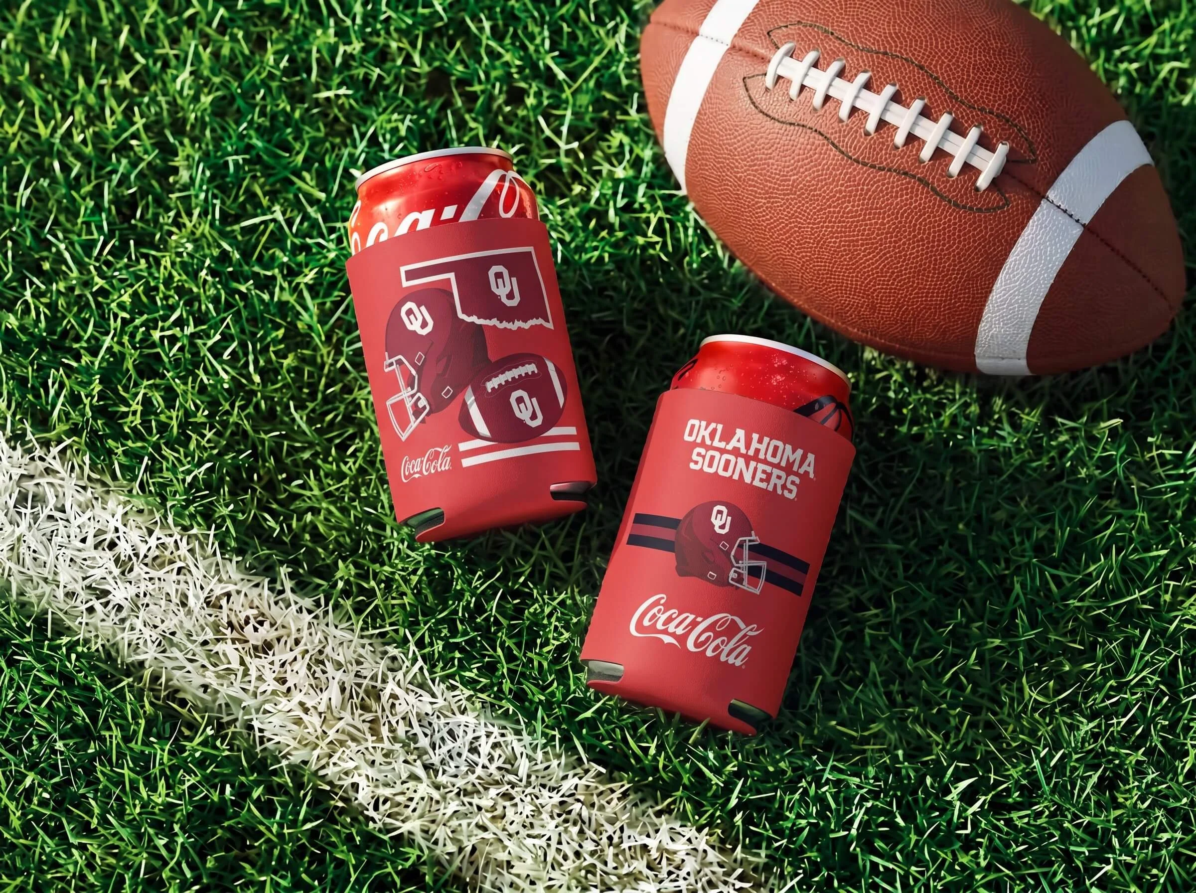 A close-up of a football on green artificial turf alongside red Coca-Cola cans with Oklahoma Sooners logos and text.
