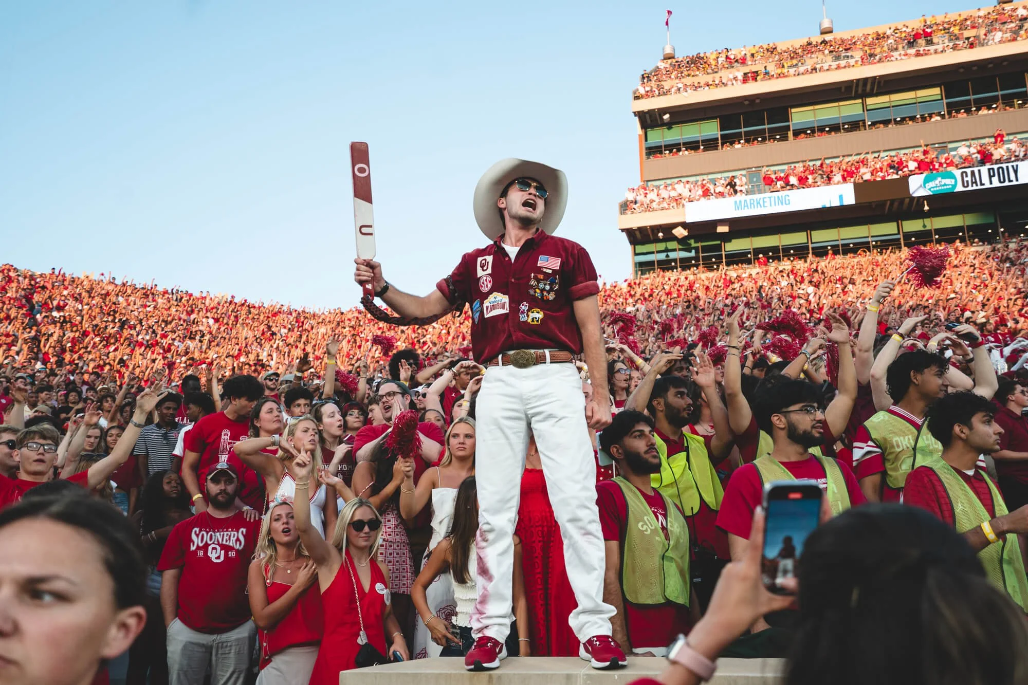 A large crowd of fans gathered at a sports stadium, many wearing red. In the foreground, a man dressed in a red Oklahoma Sooners shirt, white pants, a cowboy hat, and sunglasses stands on a platform, holding a foam finger.