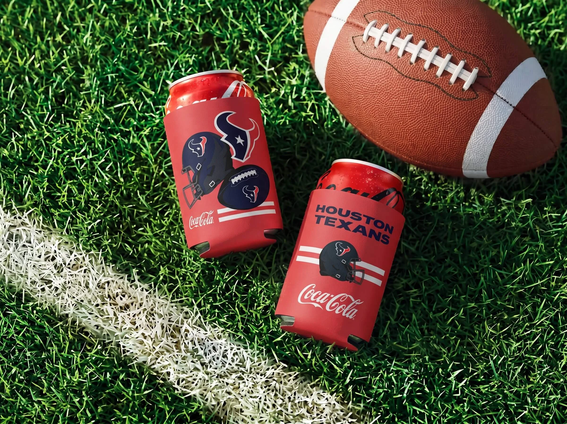 A football lying on artificial grass with white and green lines, overlapping two red soda cans with Houston Texans and Coca-Cola branding, and a brown football with white laces and white stripes.
