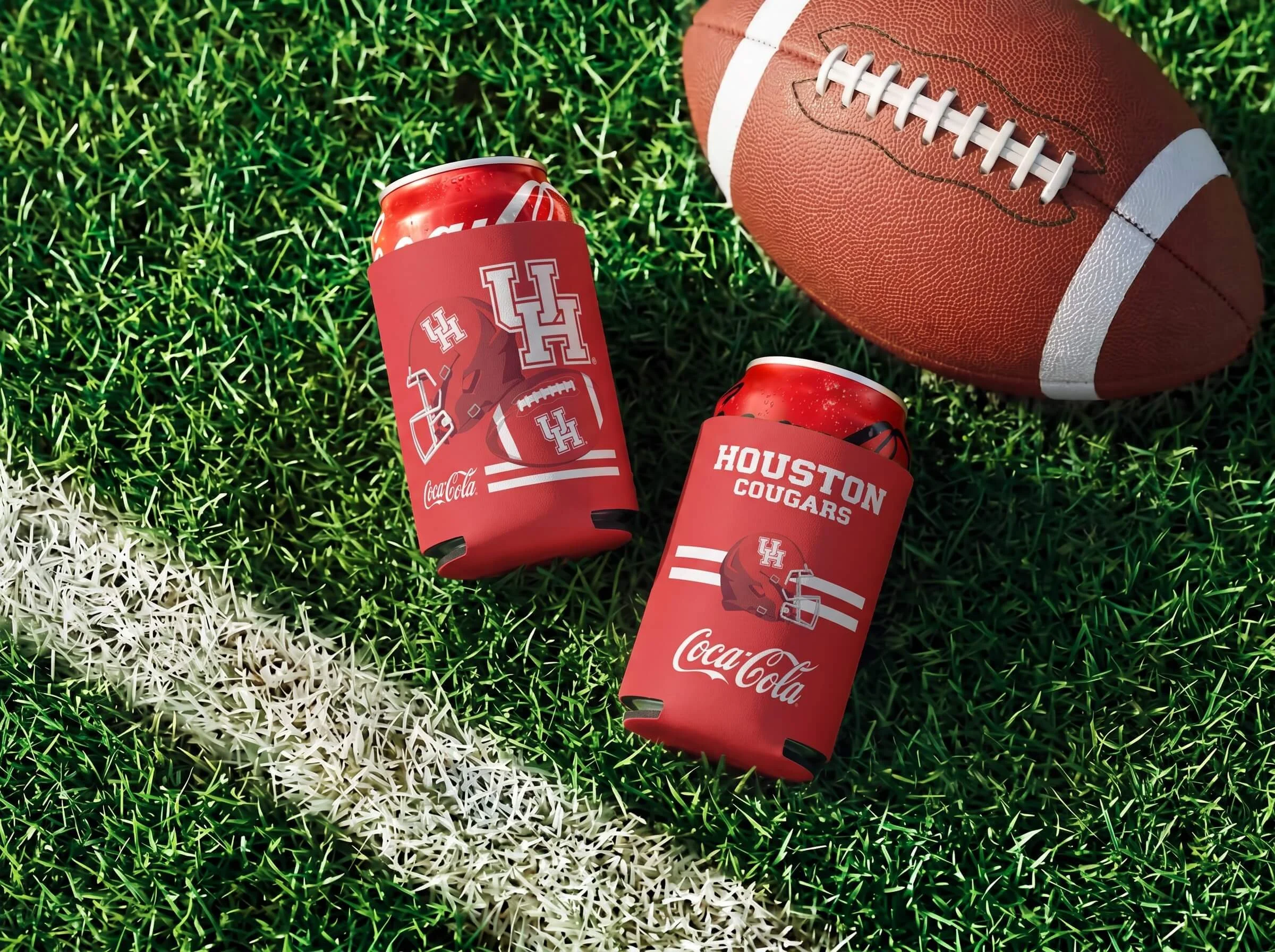 Close-up of a football and two red Coca-Cola cans with Houston Cougars logo and branding, on a grass field with white and green striped lines.