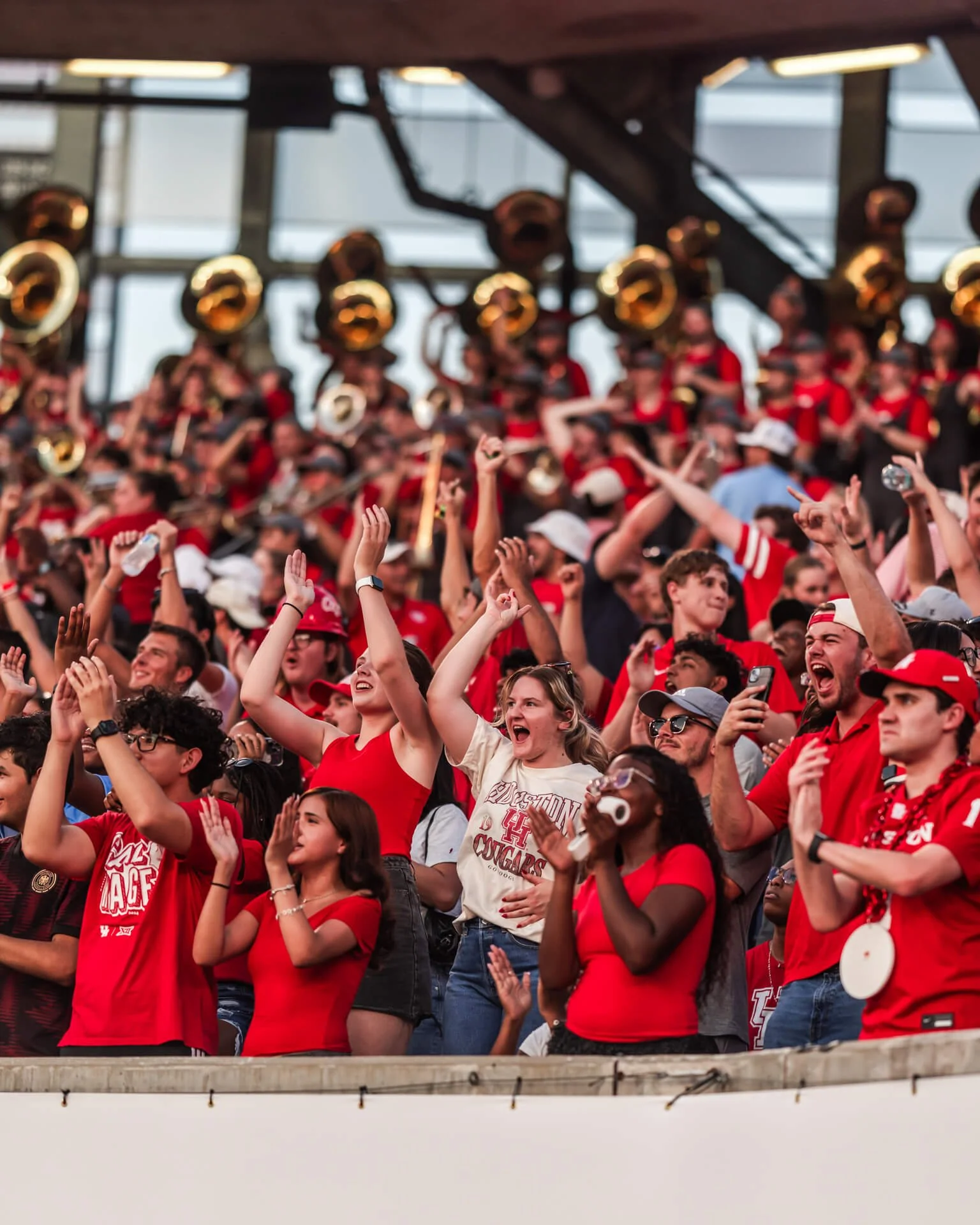 Crowd of college football fans dressed in red, cheering, with some holding up hands, in a stadium with a roof and large windows.