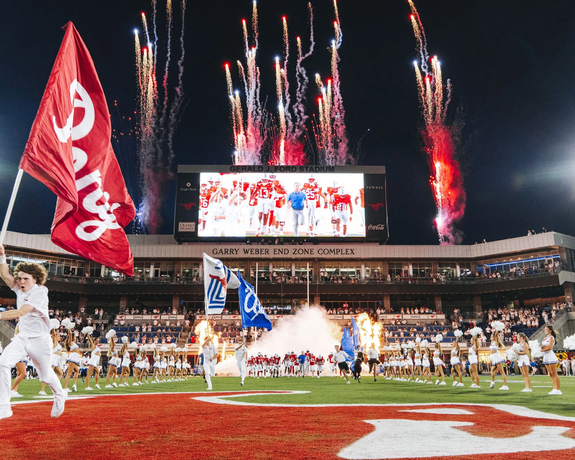 Football players and cheerleaders on the field with fireworks in the night sky at Gerald J. Ford Stadium during a game, with banners and flags in the foreground.
