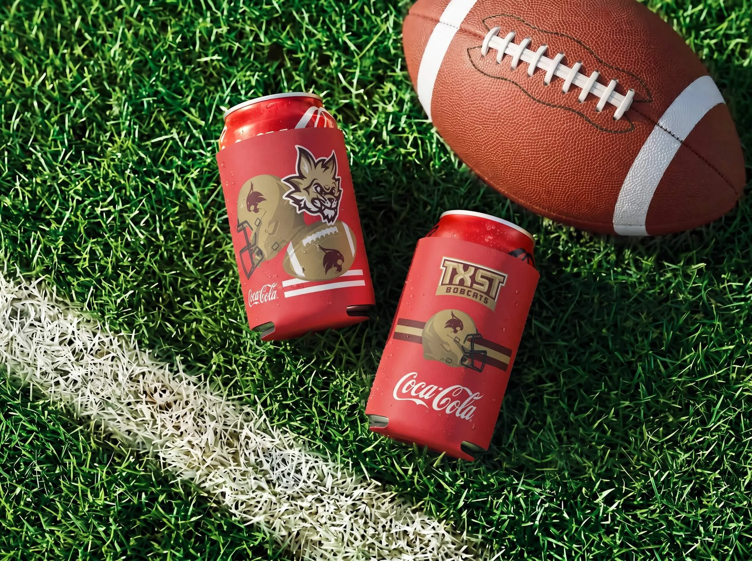 A football on a grassy field next to two cans of Coca-Cola, one wrapped in a red cozy with a university logo and the other with a title logo, with a white sideline in the grass.