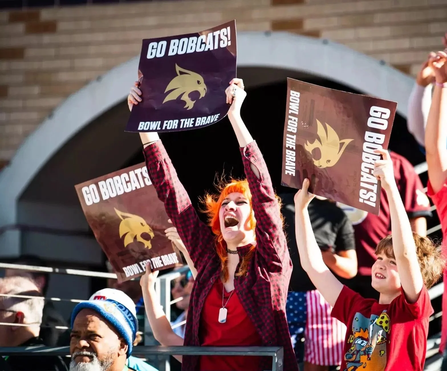 Crowd of sports fans at a stadium holding signs that say 'Go Bobcats! Bowl for the Brave' with a bobcat logo, cheering enthusiastically.