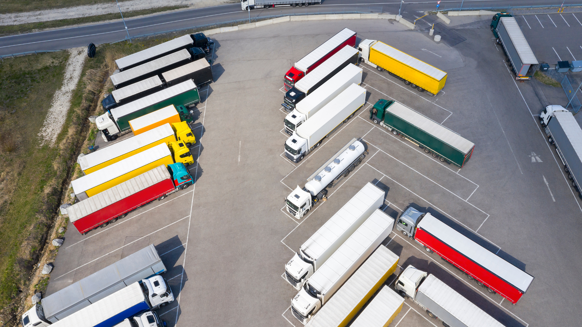 Aerial view of a parking lot filled with semi-trailer trucks in various colors including red, yellow, green, black, and white parked in designated spaces.