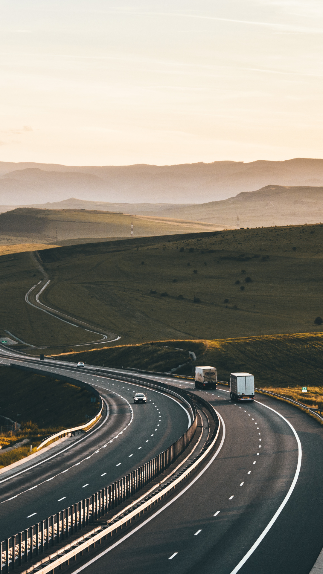 A winding highway with a few trucks and cars driving through green rolling hills at sunset or sunrise.