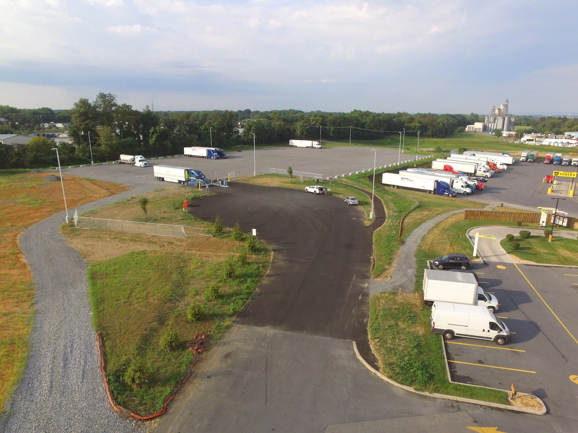An aerial view of a parking lot with several trucks, cars, and a small building with a sign that says 'CERTIFIED SCALES.' There are green trees and a grain elevator in the background, with a blue sky and some clouds.