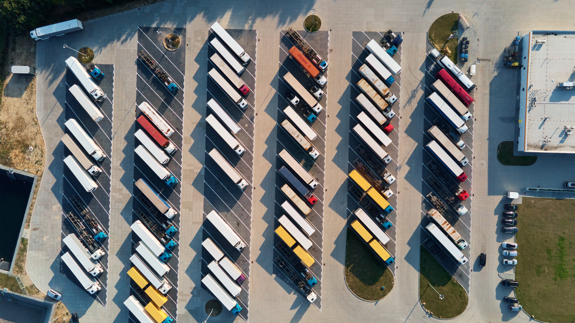 Aerial view of a parking lot filled with trucks and trailers parked in rows, with some small patches of grass and trees around.