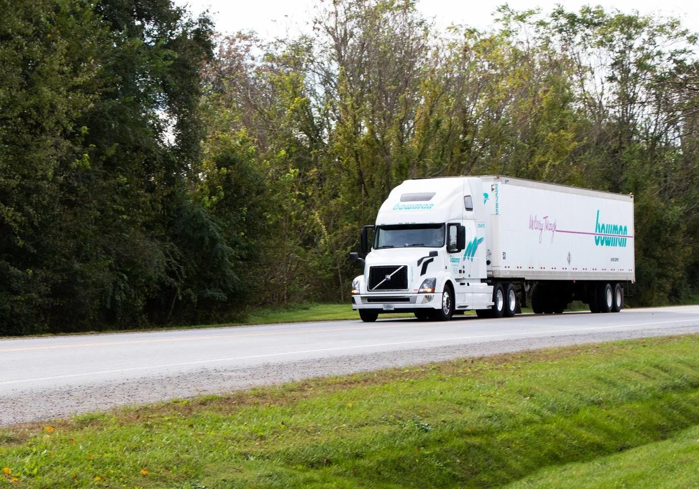 A large white semi-truck driving on a highway with a grassy shoulder and trees in the background.