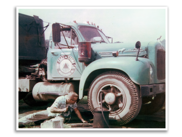An older man working on a large, vintage truck with a faded blue-green color, parked outdoors. The truck has a logo on the door that reads "The CON-STOP" and features an icon of a traffic cone. The man is lying on the ground and using tools under the truck.