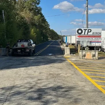 A two-lane road with a pickup truck parked on the left side and a large OTP Byron truck on the right side near a small parking area with yellow-striped spaces. Trees line the left side, and utility poles run along the road under a partly cloudy sky.
