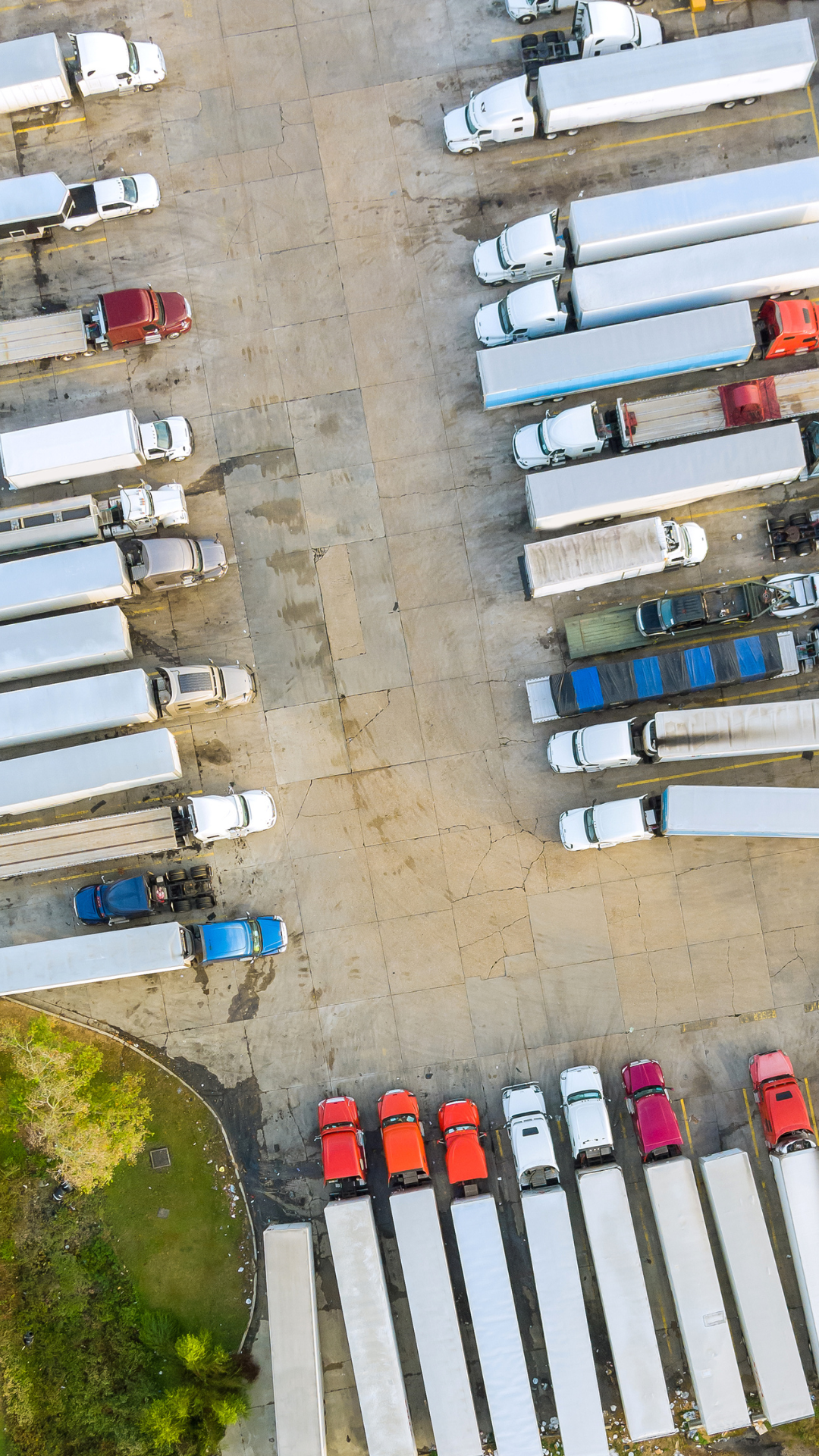 Aerial view of a parking lot with white trucks and a few cars parked, and a small green landscaped area with trees at the bottom left corner.